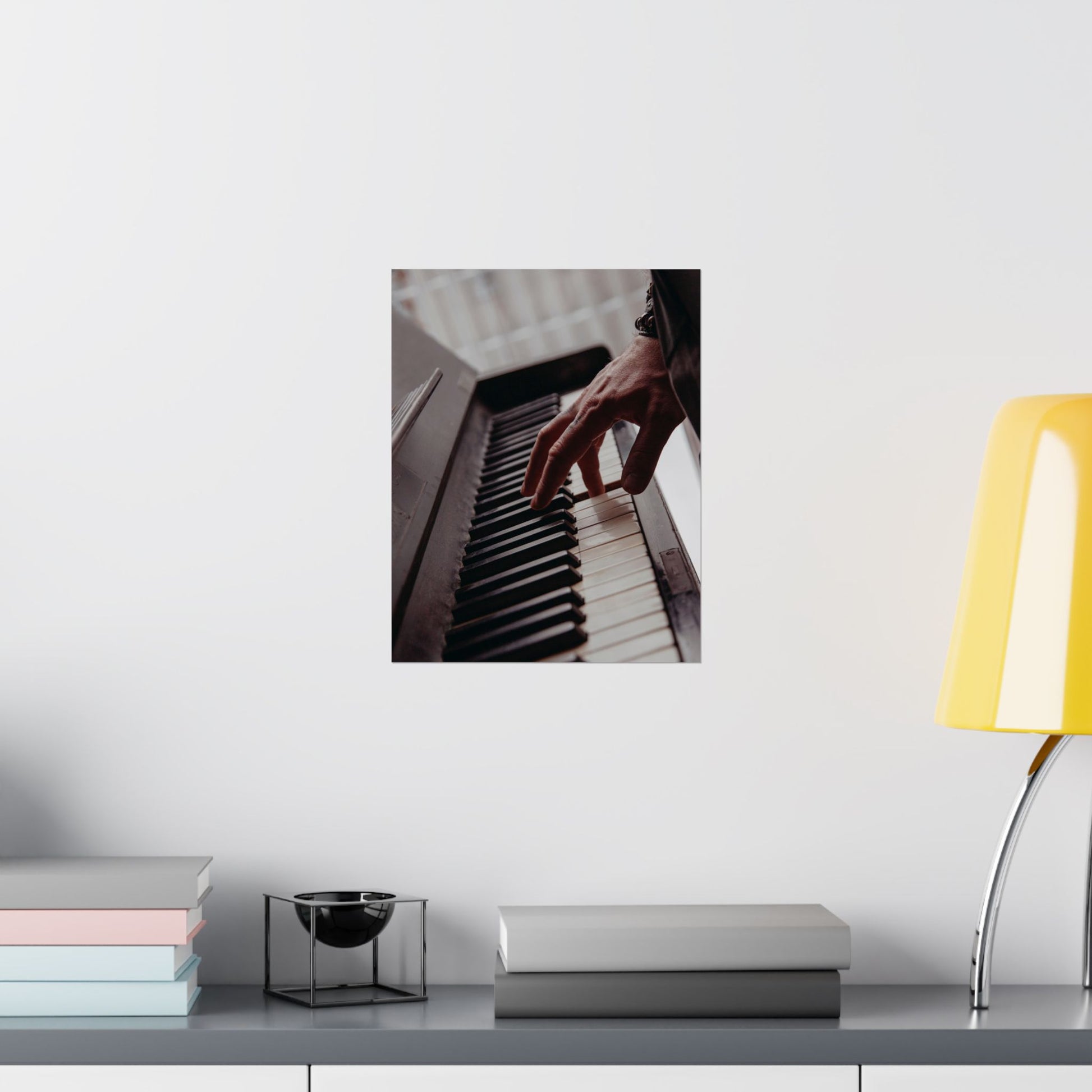 Framed picture of hands playing piano on a wall above a desk with books and a lamp.