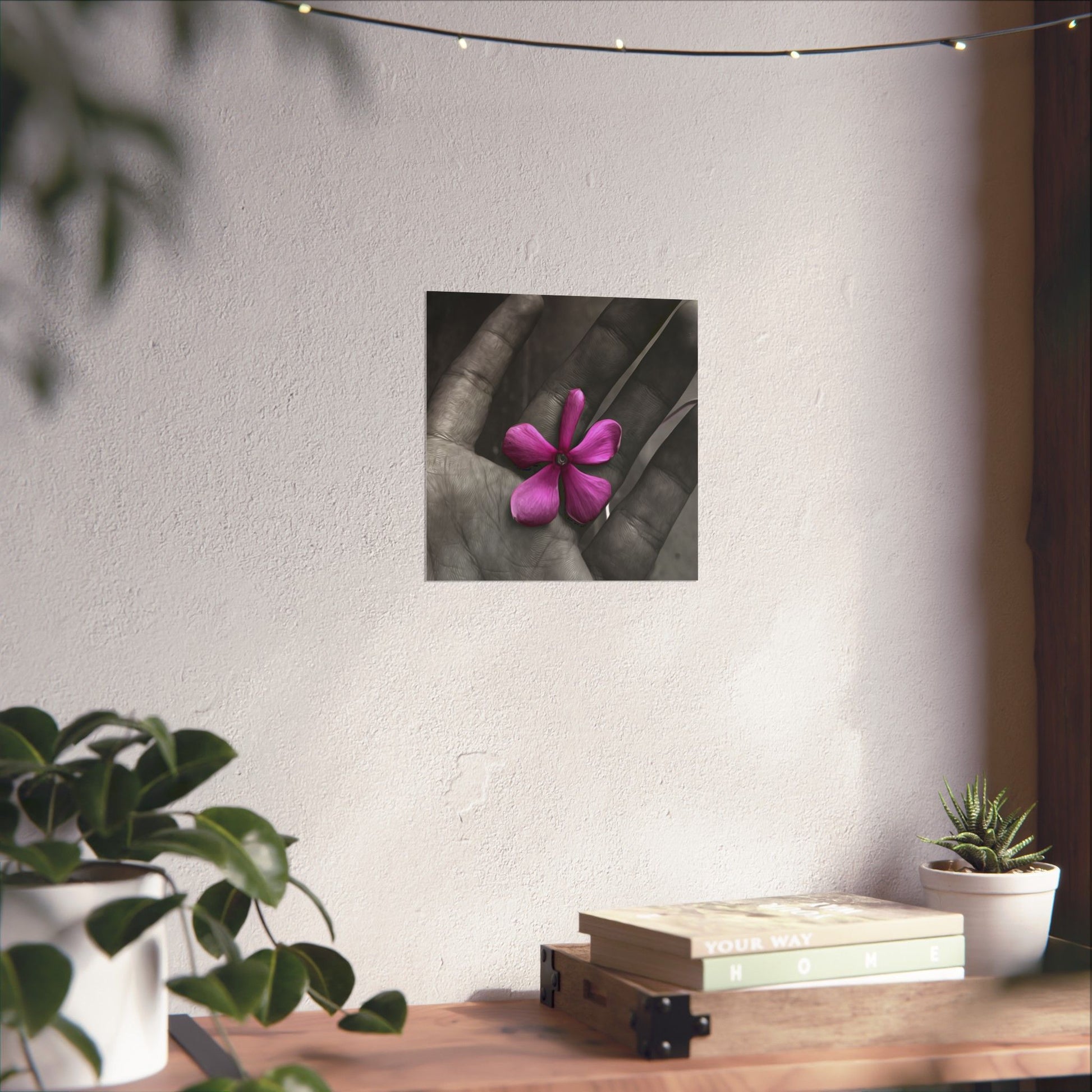 Framed artwork of a pink flower held in a hand on a wall above a wooden desk with plants.