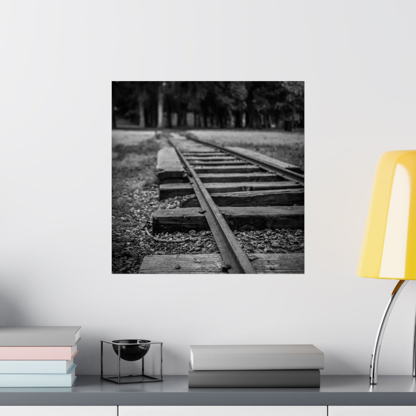 Black and white photograph of a railway track on a wall above a shelf with books and a lamp.