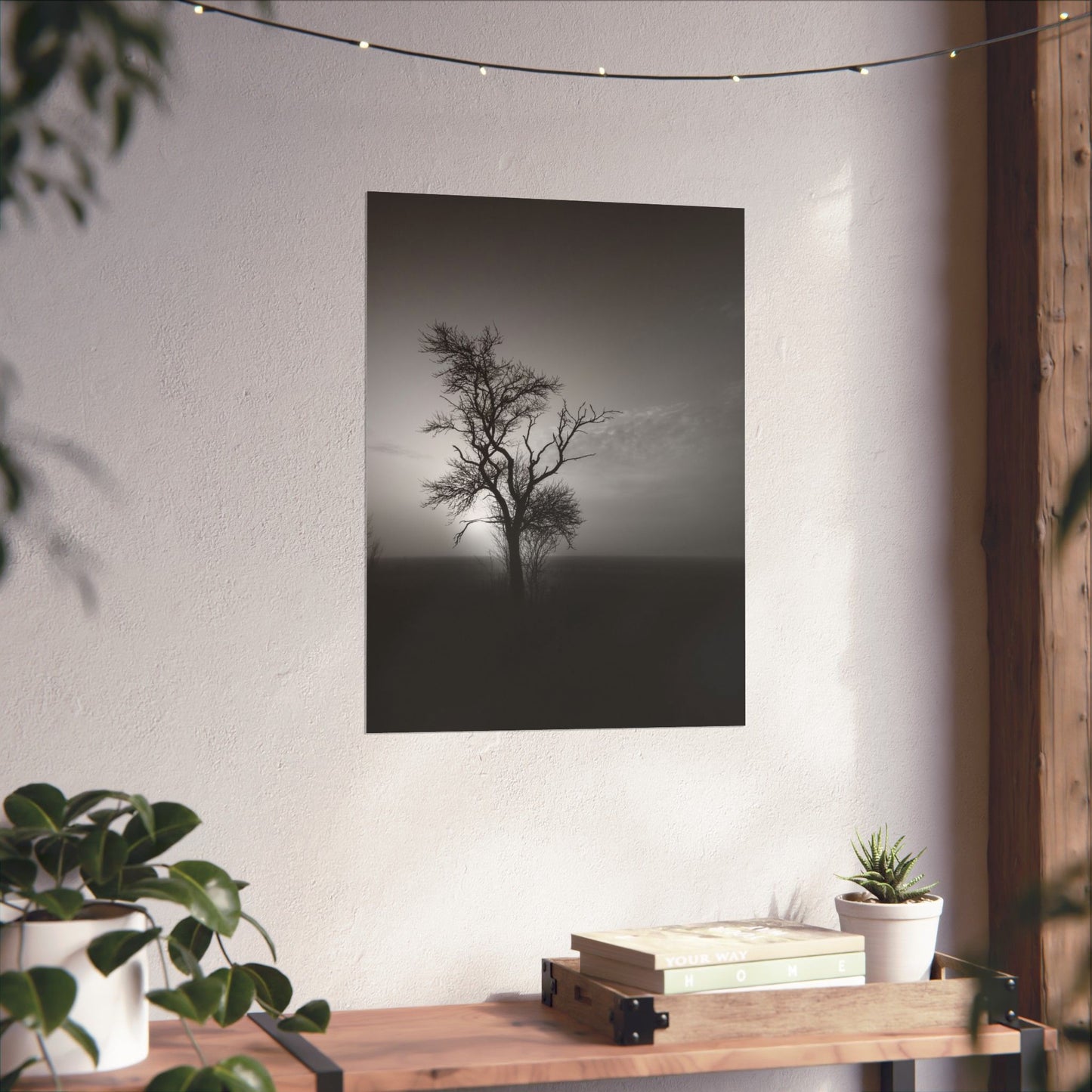 Art print of a lone tree on a hillside hanging on a white wall above a wooden table with plants.