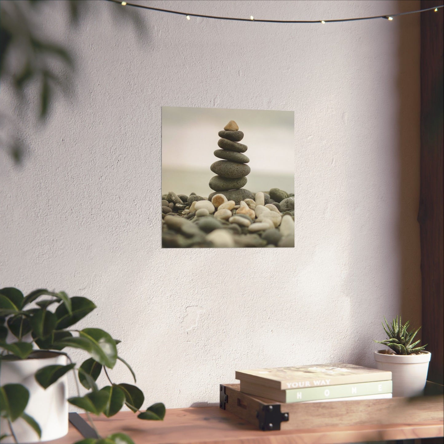 Wall art of stacked stones on a white wall with plants and books below.