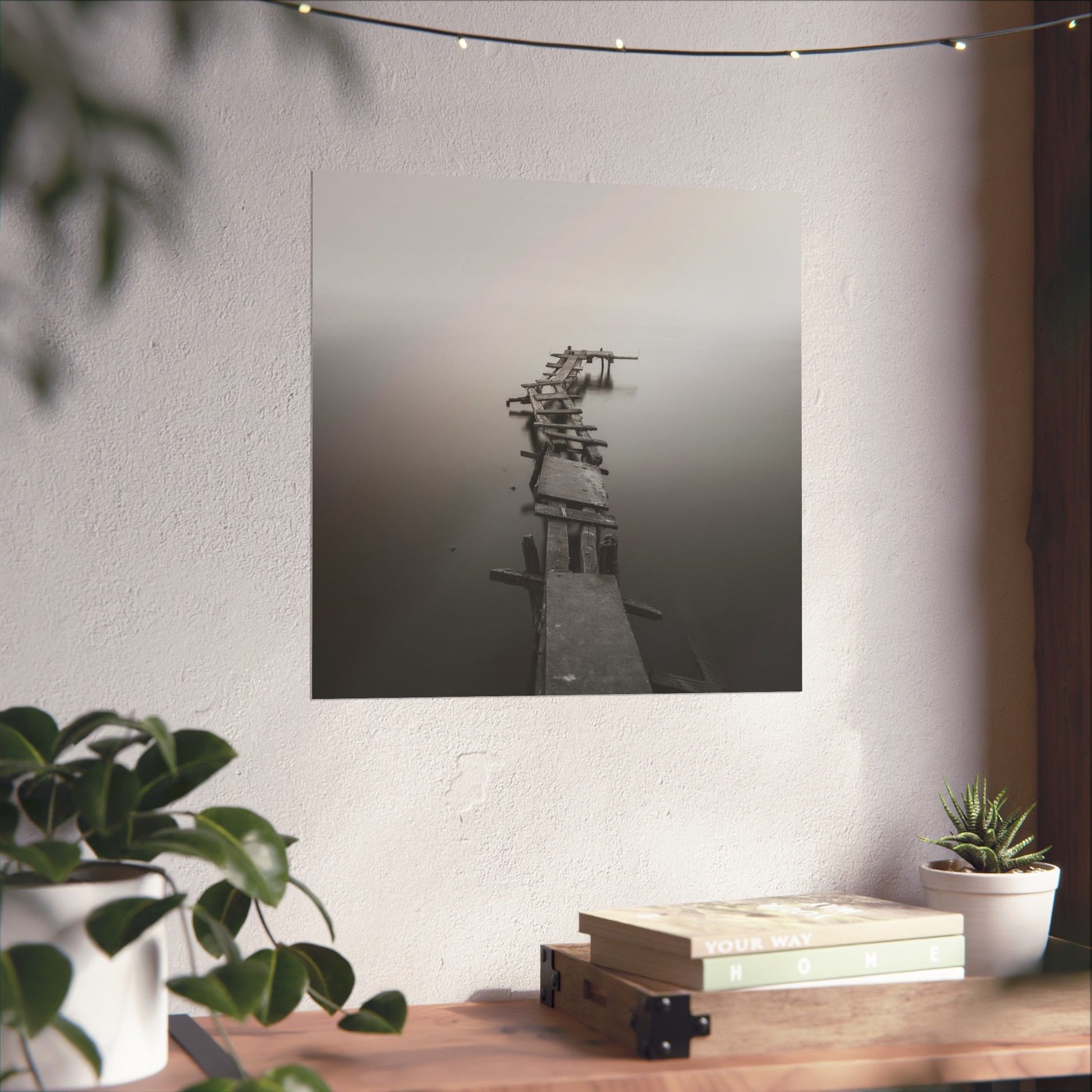 Framed black and white photograph of a dock on a wall above a wooden desk with plants and books.
