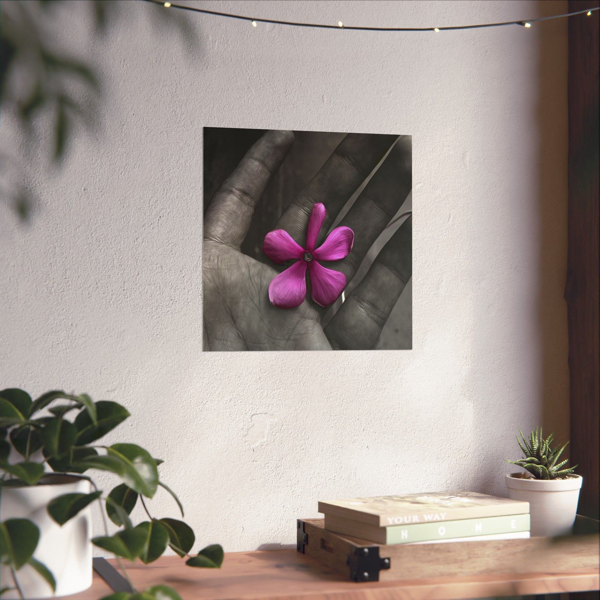 Framed artwork of a hand holding a pink flower on a wall above a wooden table with plants.