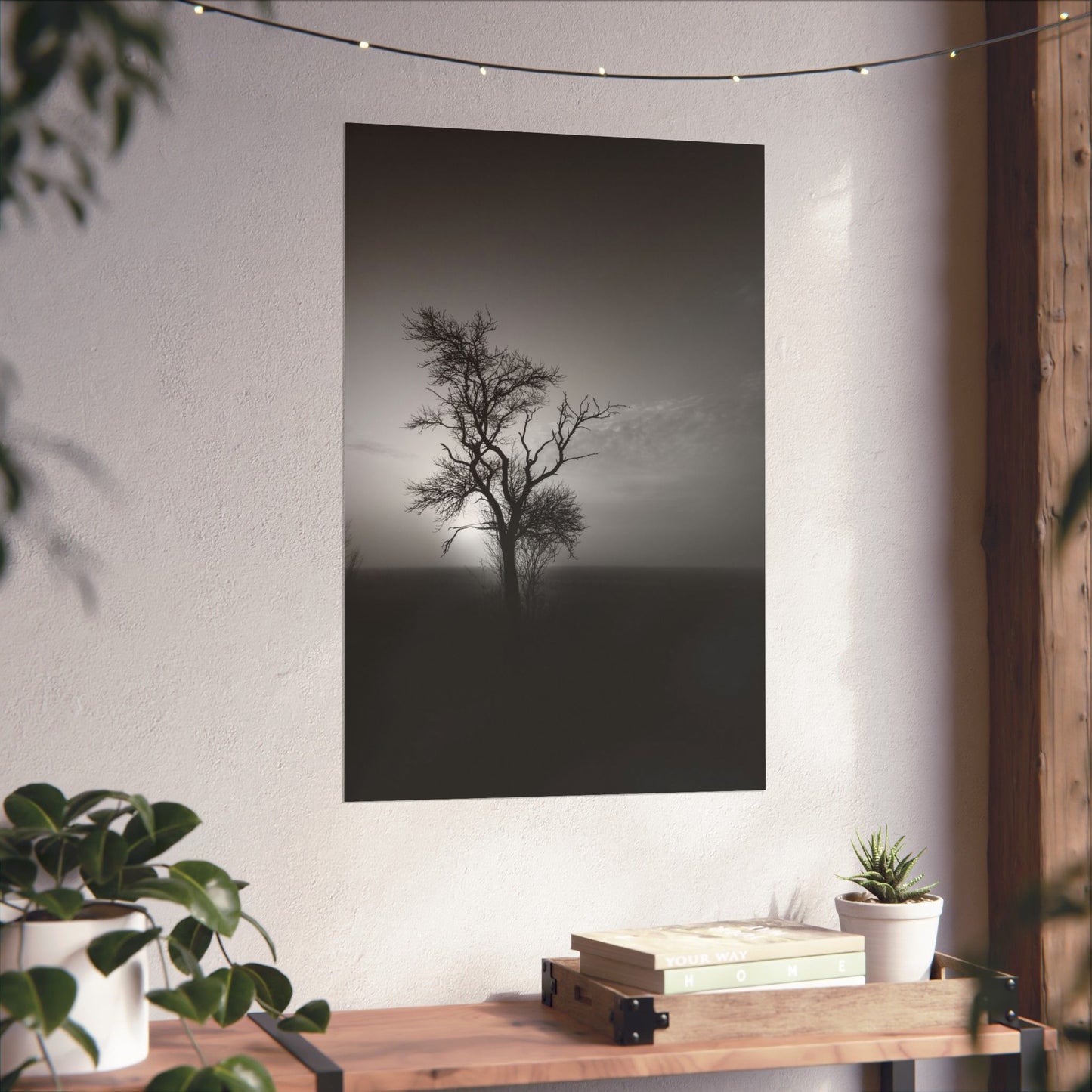 Art print of a lone tree on a wall in a home setting with plants and books.