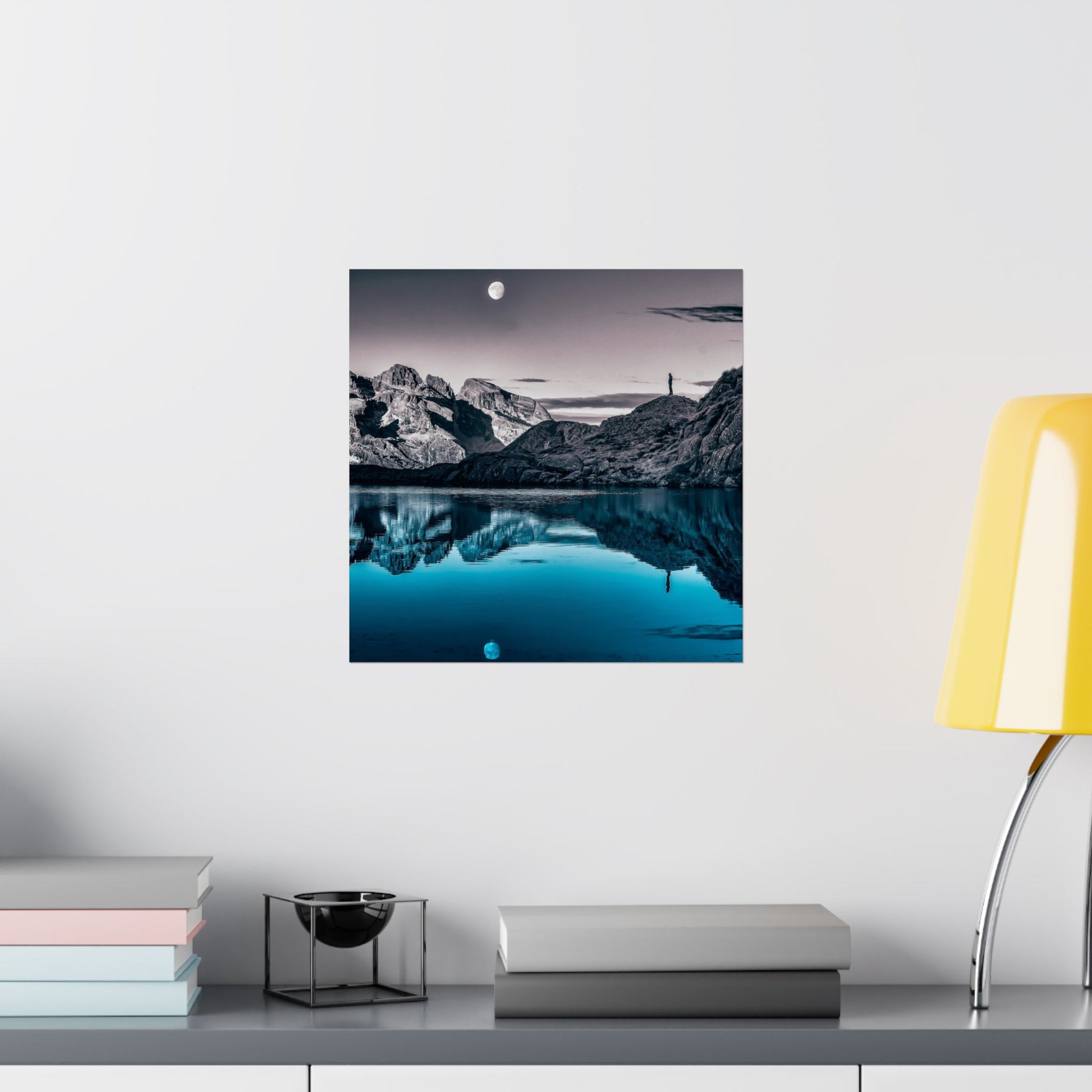 Framed landscape print of a mountainous landscape with a lake on a white wall above a shelf.