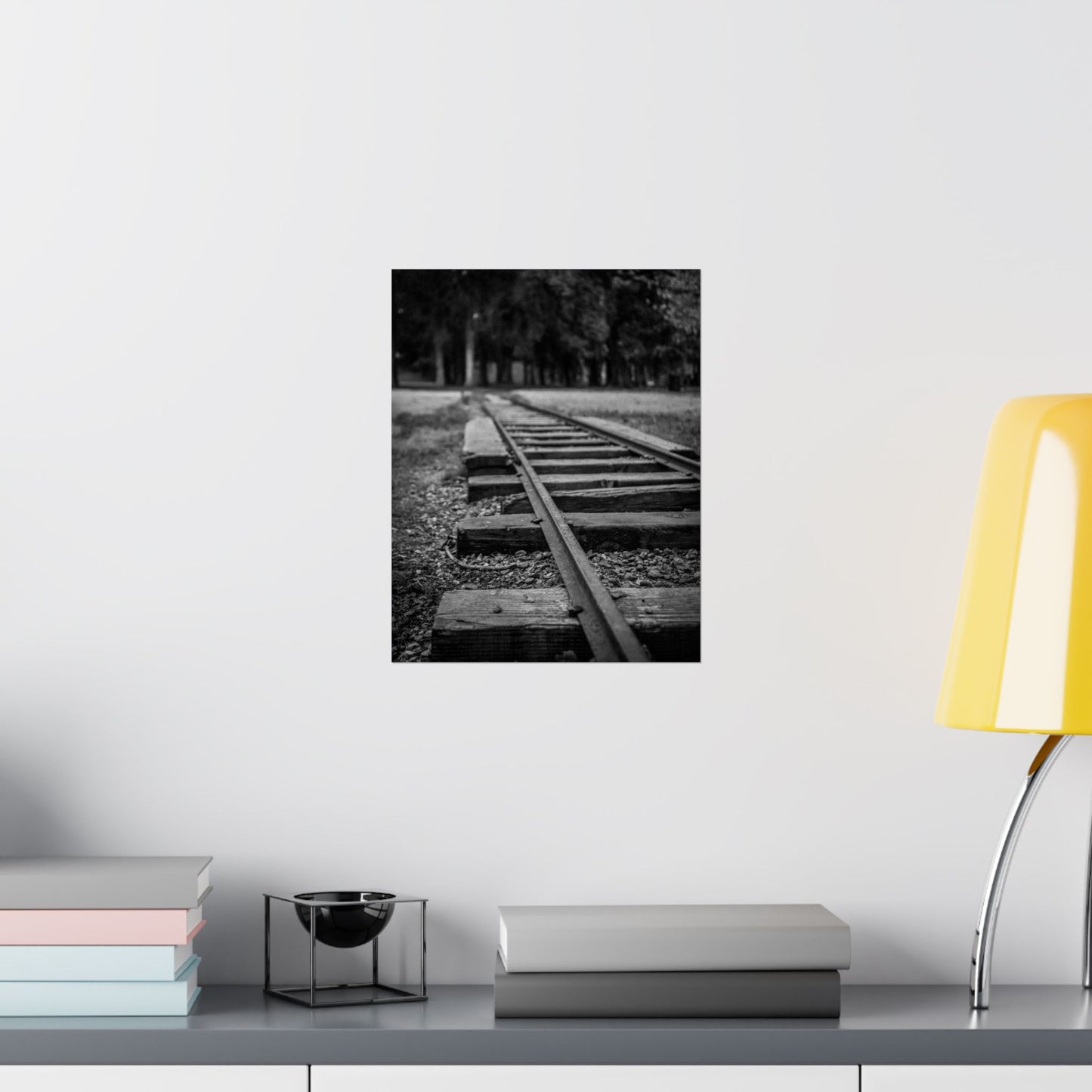Black and white photograph of a railway track on a wall above a shelf with books and a lamp.
