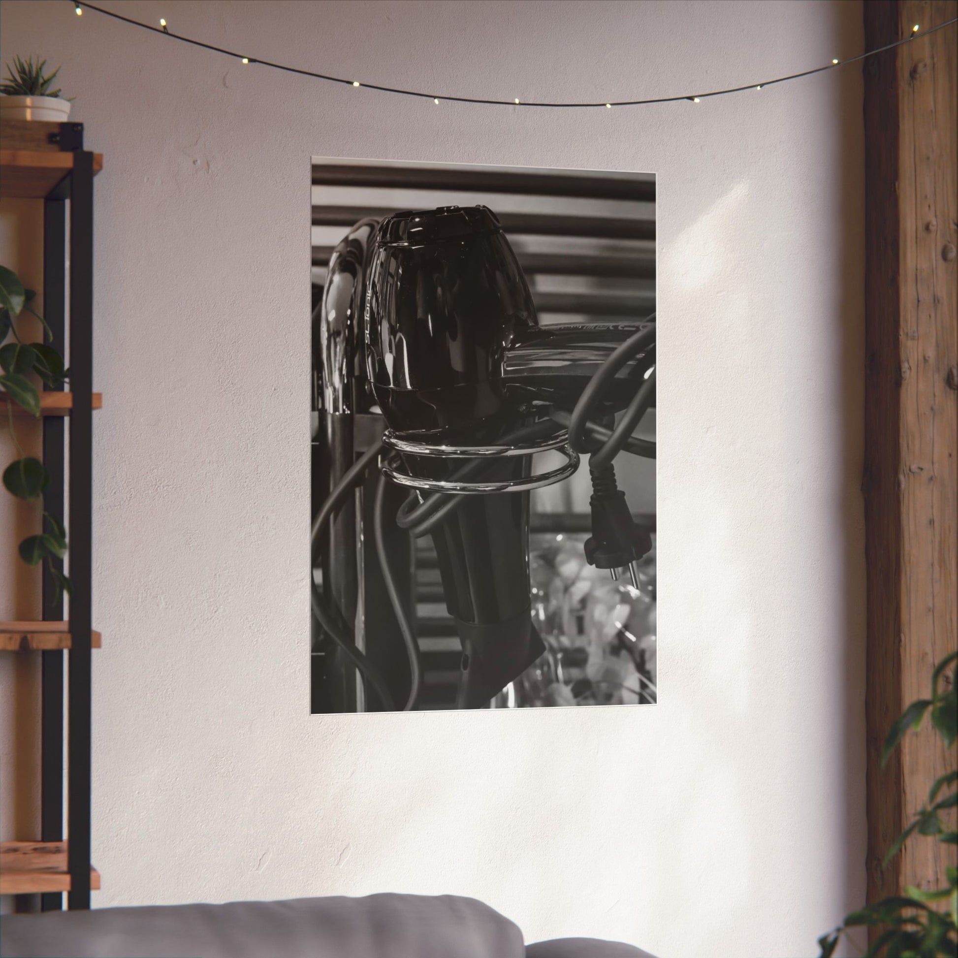 Black and white photograph of a vase on a reflective surface, framed by string lights and a plant.