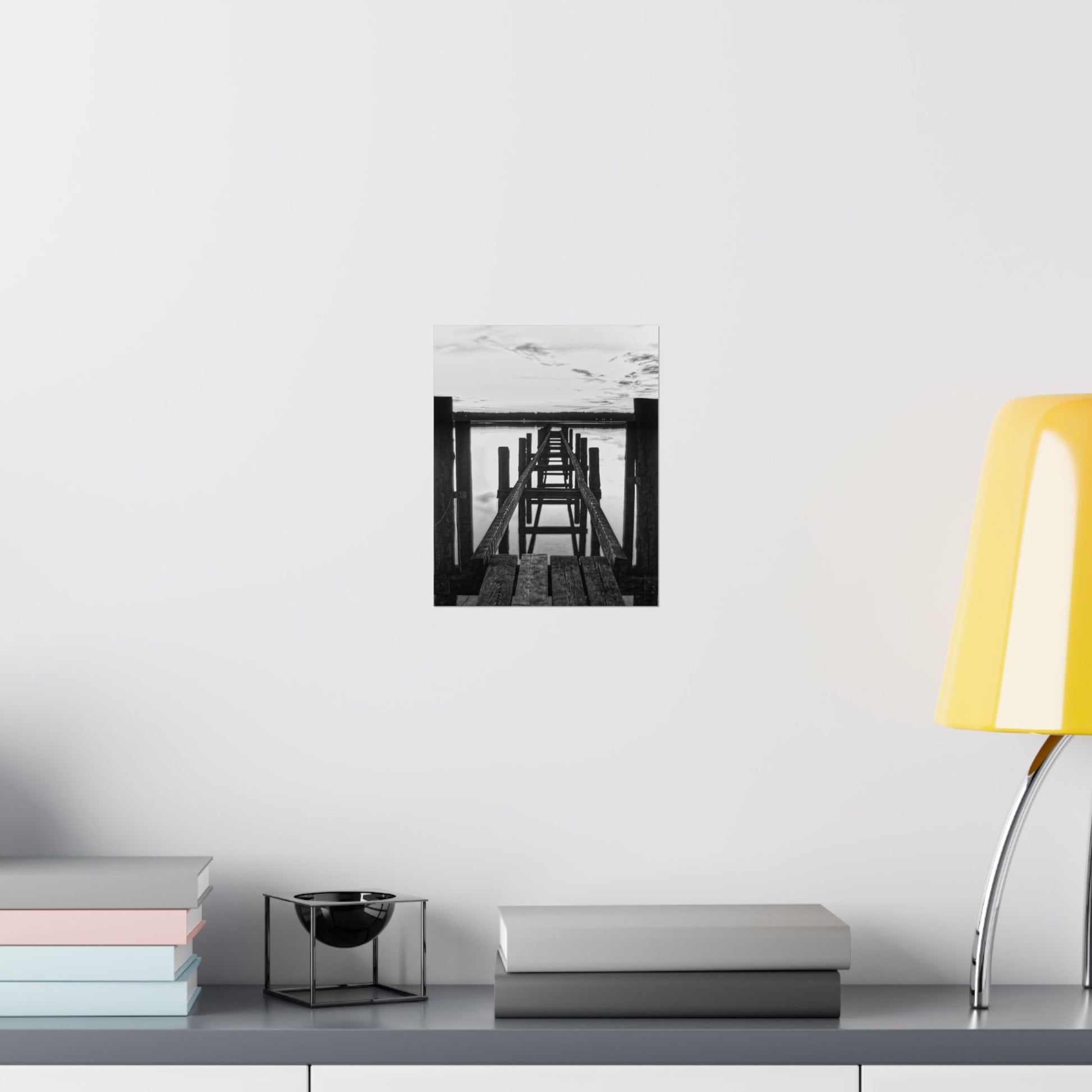 Framed black and white artwork on a wall above a desk with books and a lamp.
