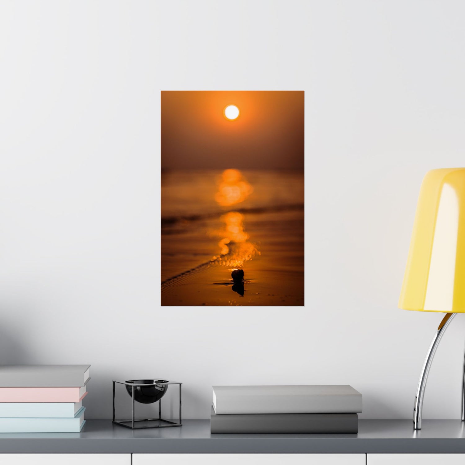 Framed artwork of a sunset over water on a wall above a desk with books and a lamp.