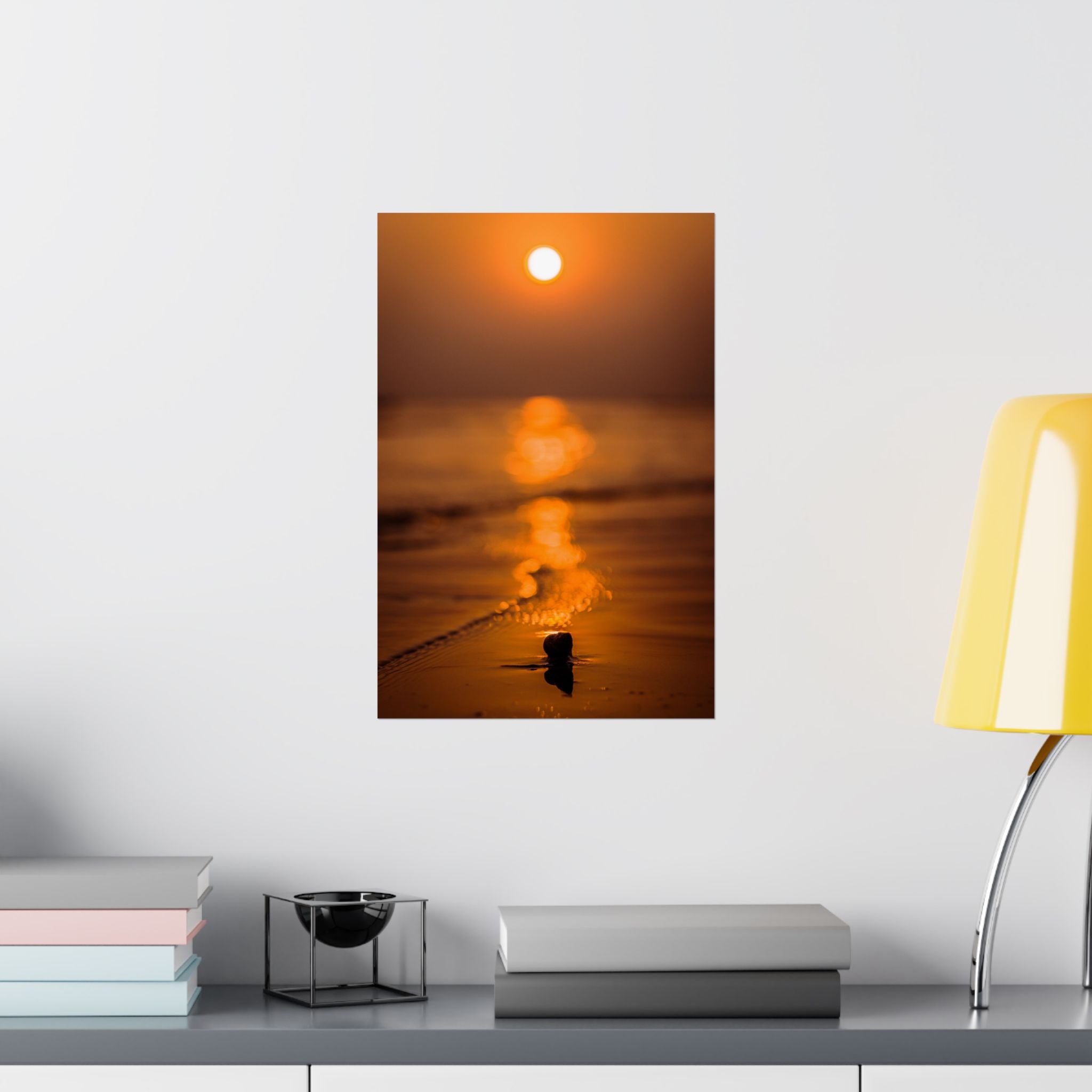 Framed artwork of a sunset over water on a wall above a desk with books and a lamp.