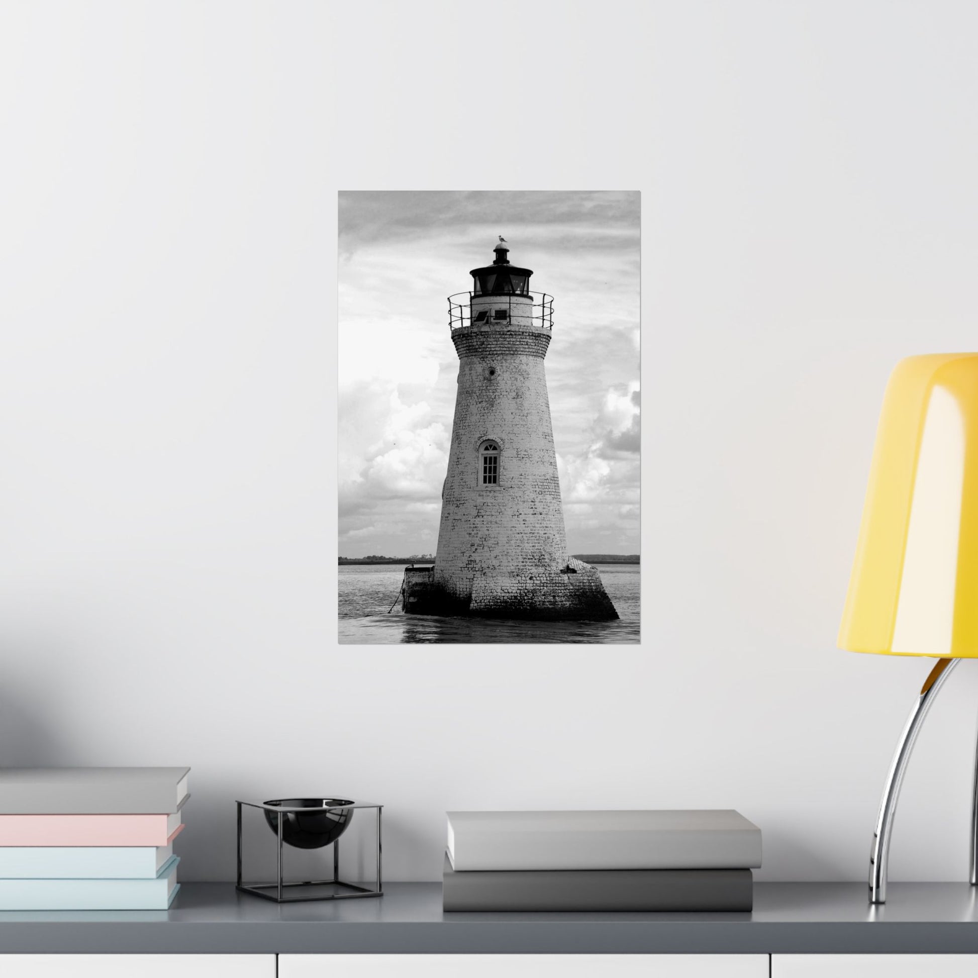 Poster of a lighthouse on a wall with books and a lamp on a table below.