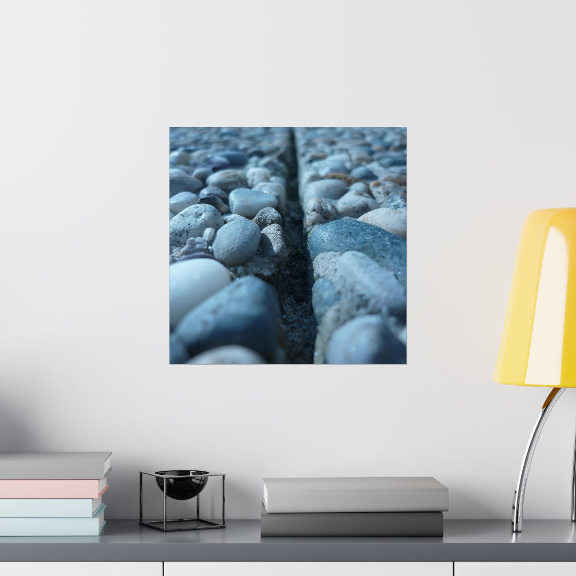 Framed artwork of pebbles on a wall above a shelf with books and a lamp.
