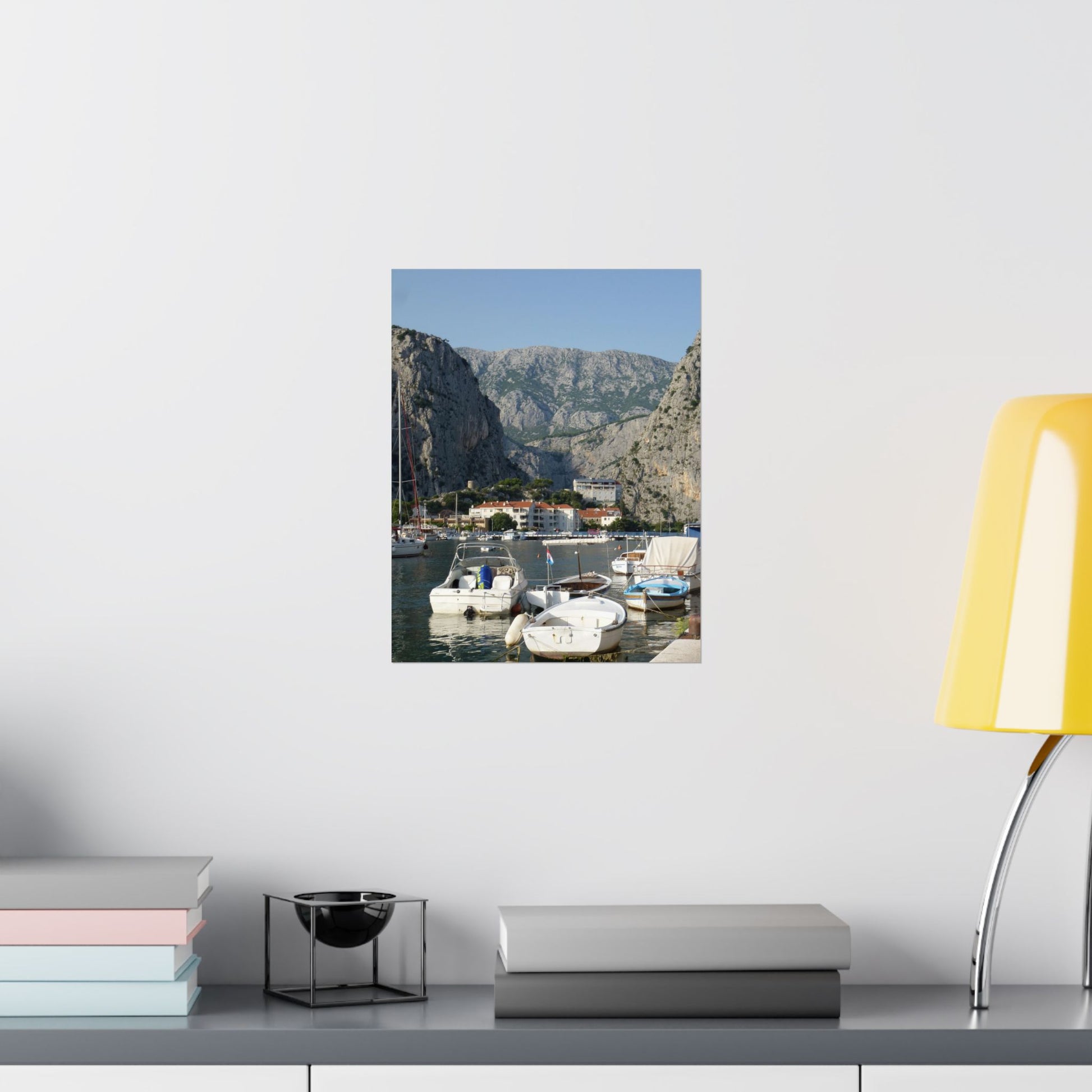 Scenic wall art of a coastal village with mountains on a white wall, next to books and a lamp.