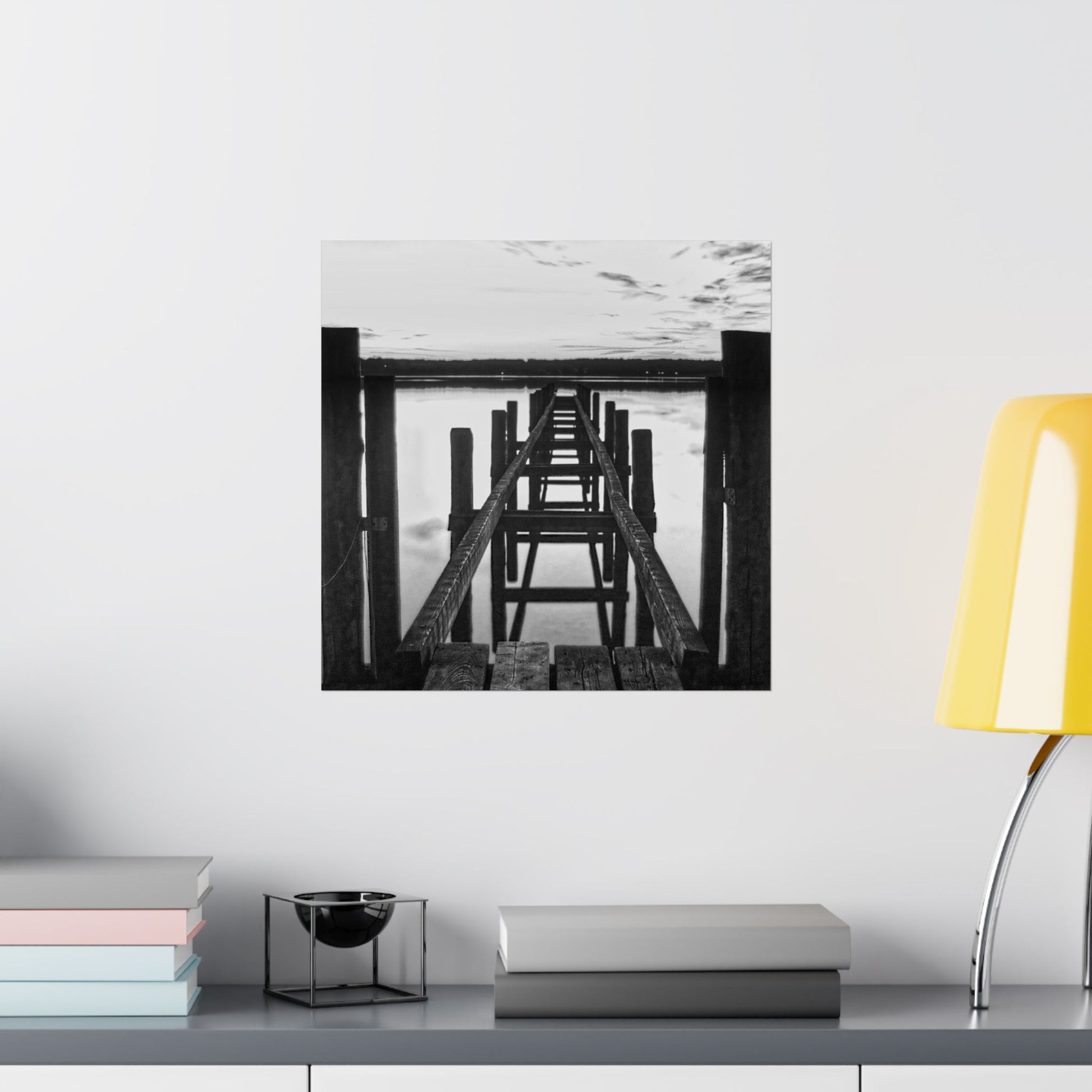Black and white photo of a pier on a wall above a shelf with books and a lamp.