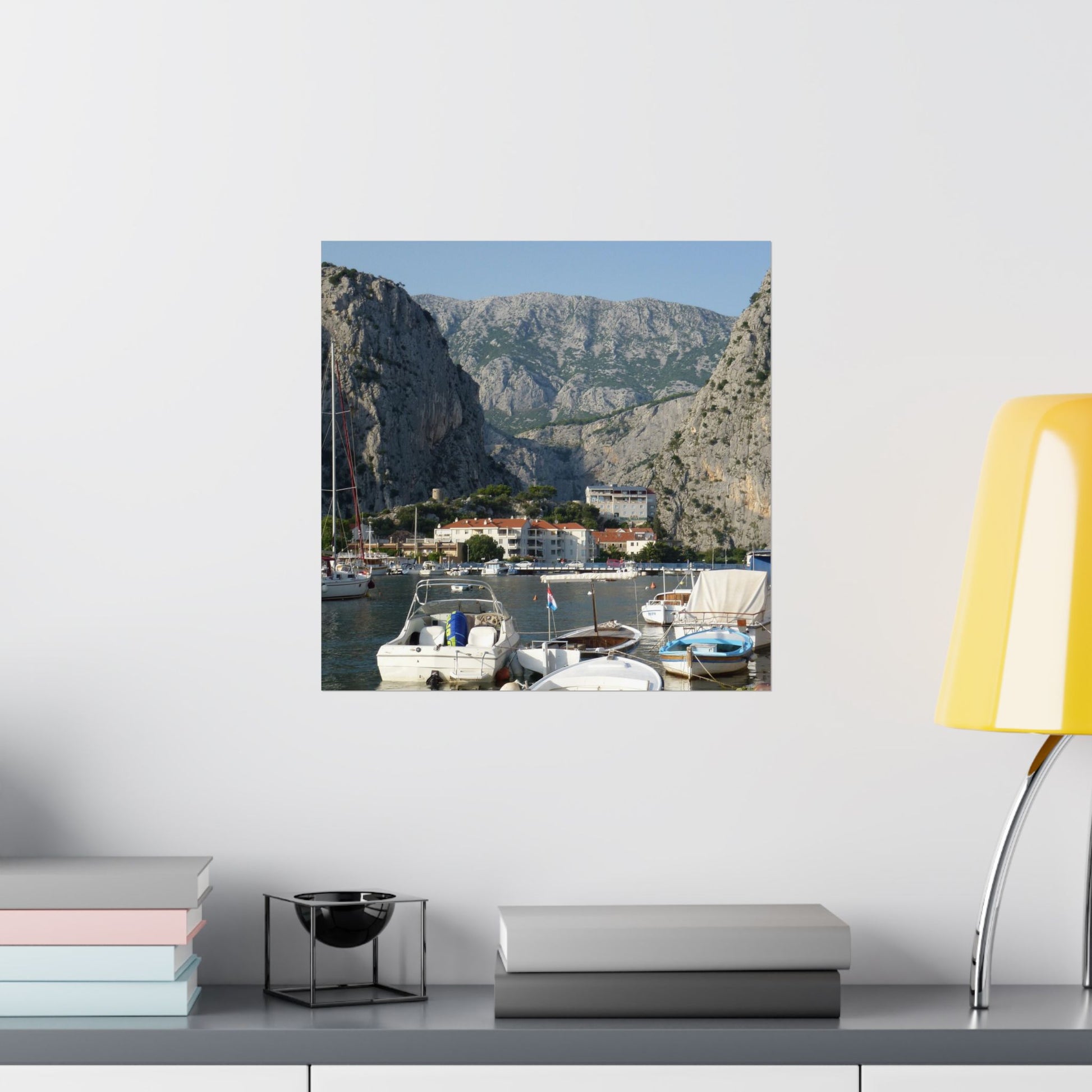 Canvas print of a coastal scene with boats and mountains on a wall above a shelf.