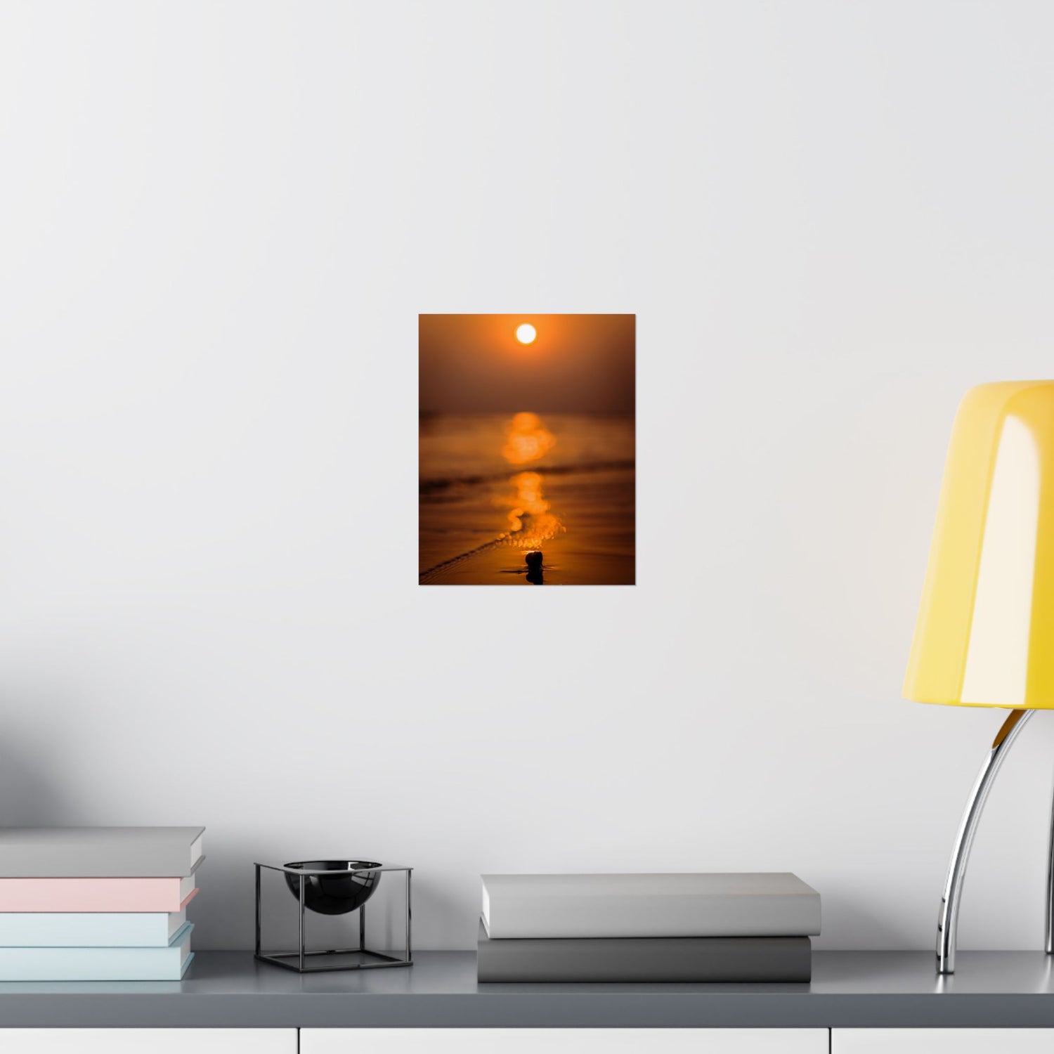 Wall art of a sunset over water on a white wall above a shelf with books and a lamp.