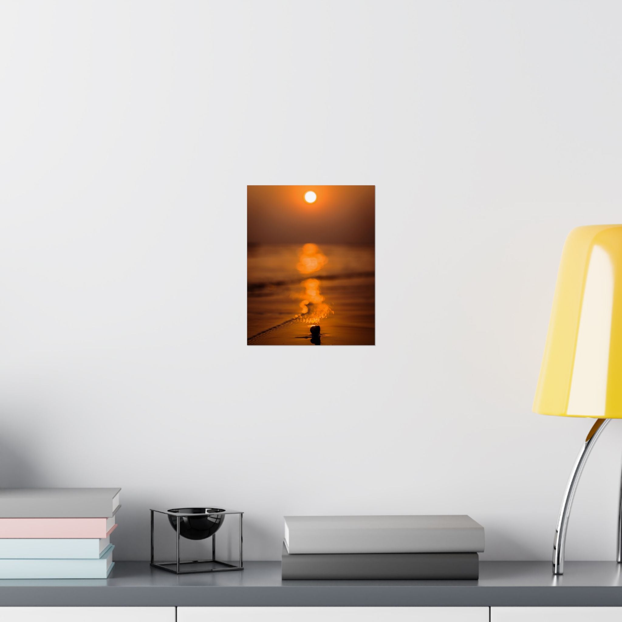 Wall art of a sunset over water on a white wall above a shelf with books and a lamp.