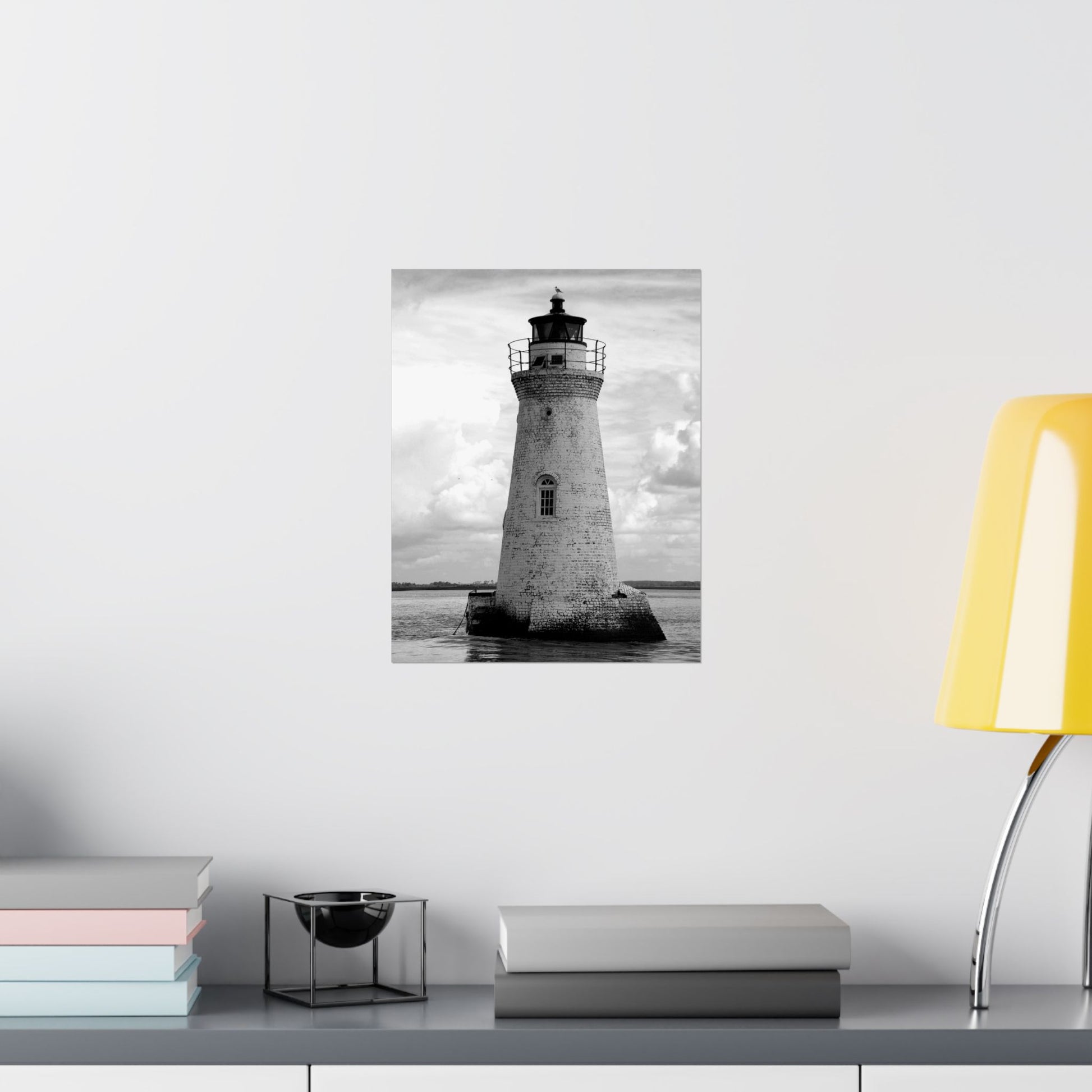Poster of a lighthouse on a wall with books and a lamp on a table below.