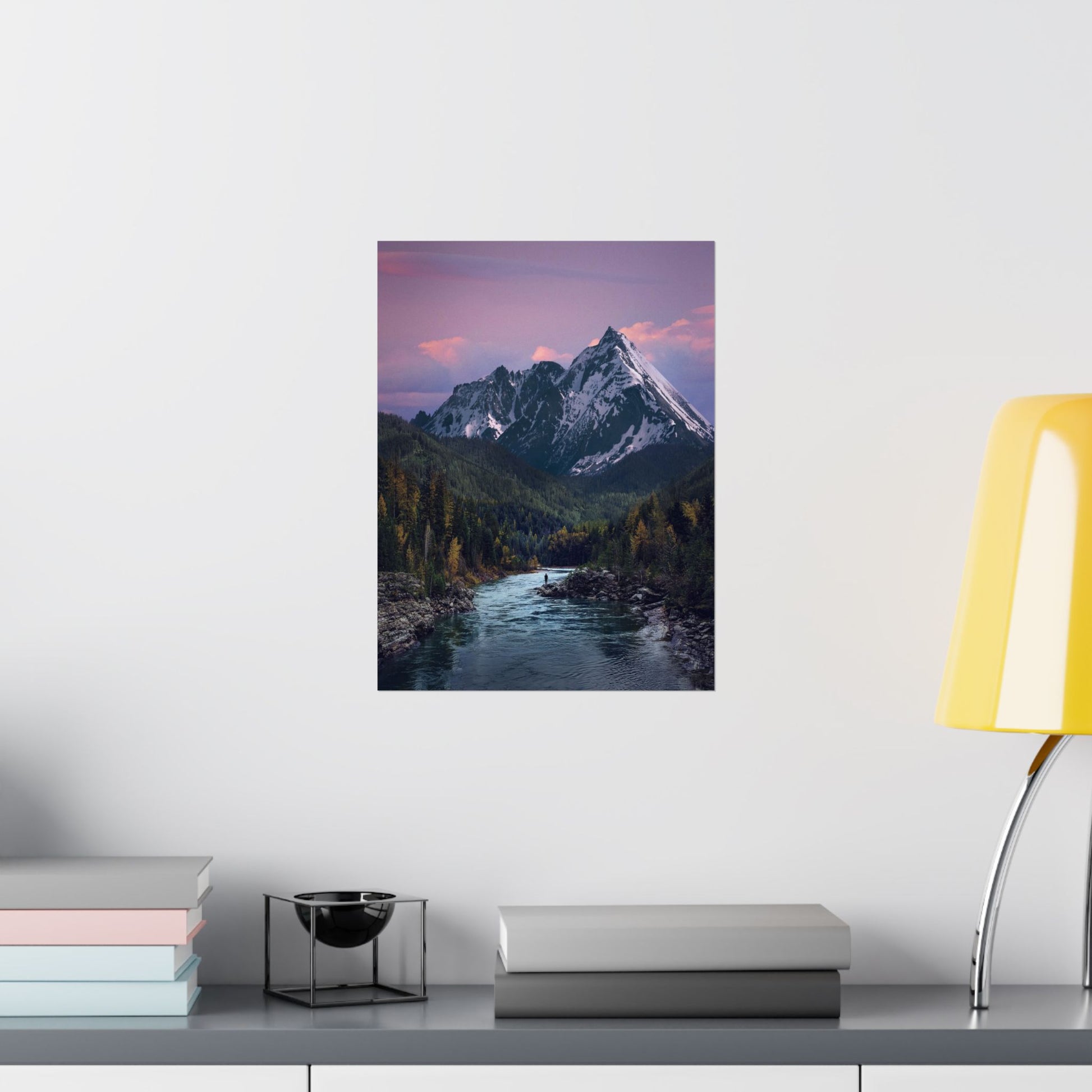 Framed mountain landscape print on a wall above a desk with books and a lamp.