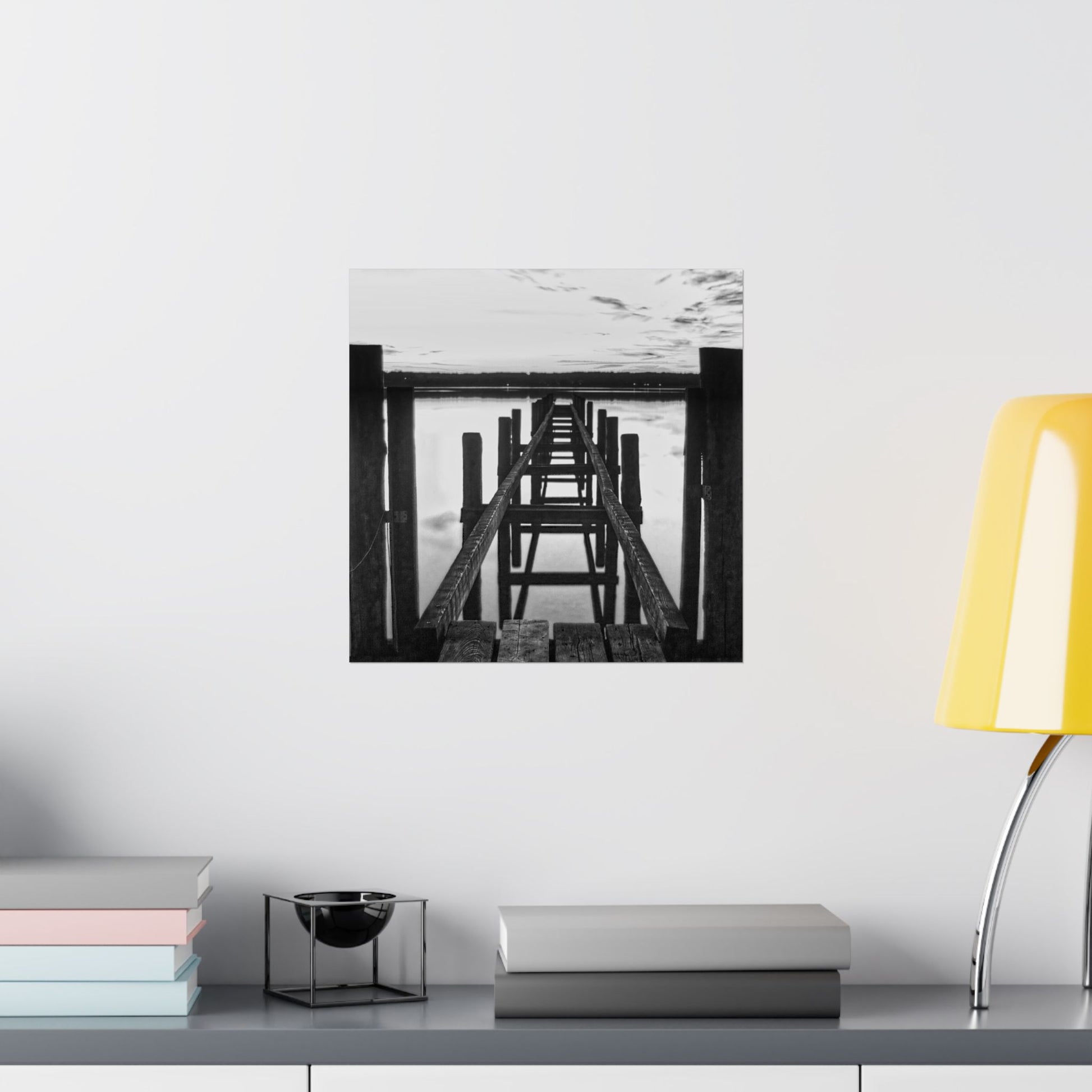 Framed black and white artwork on a wall above a shelf with books and a lamp.