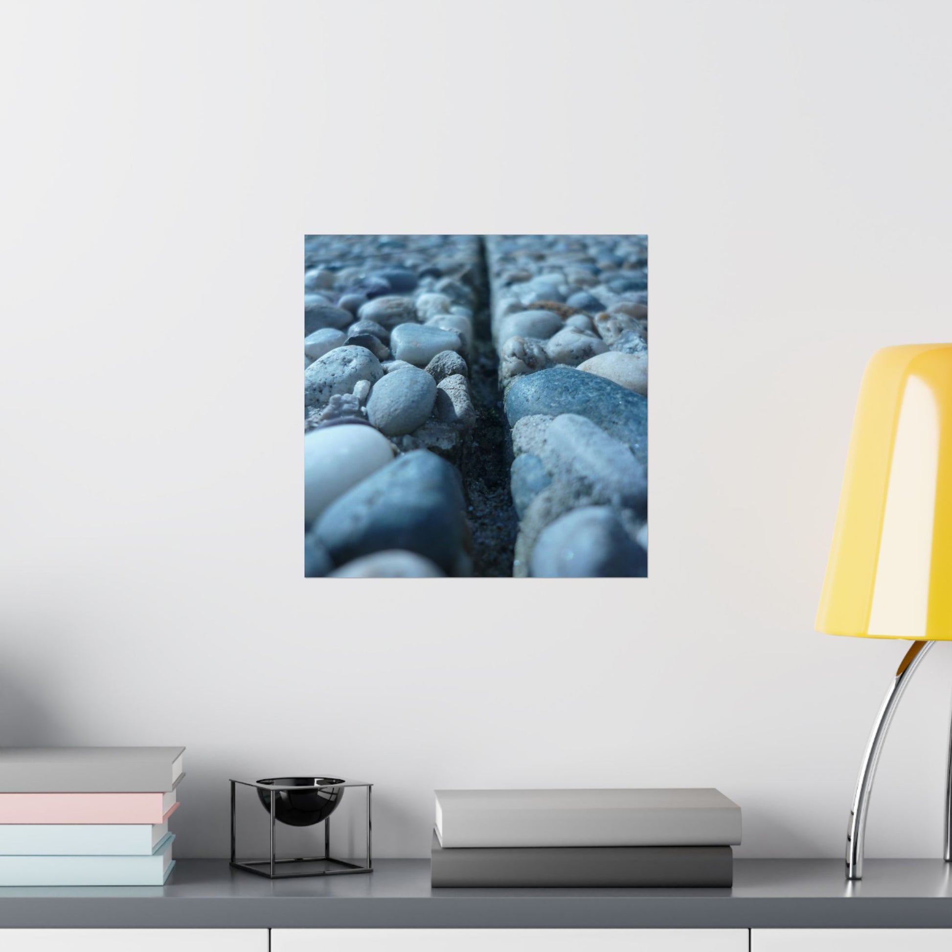 Framed artwork of pebbles on a wall above a shelf with books and a lamp.