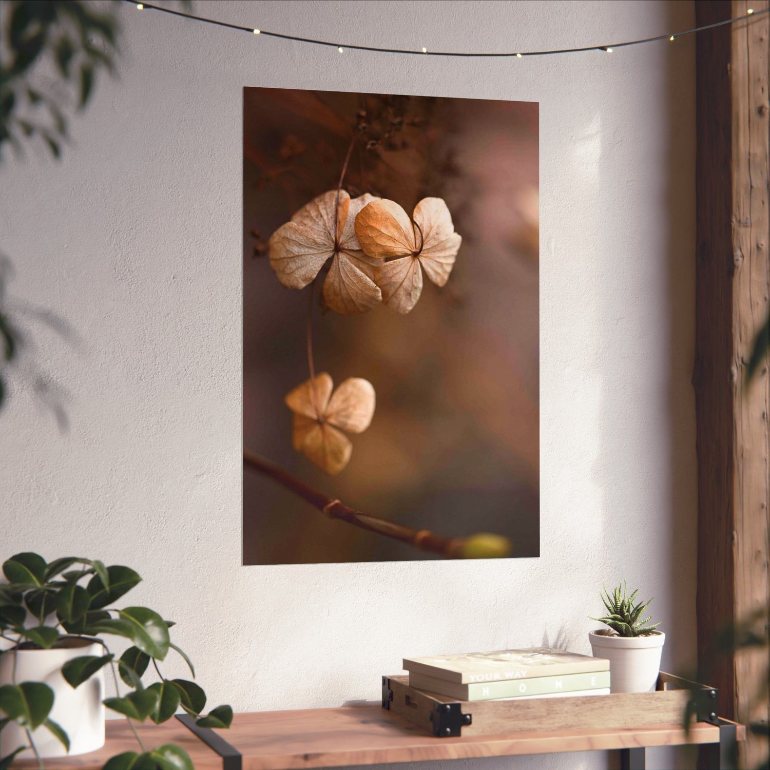 Framed artwork of dried flowers on a wall with a wooden table and plants below.
