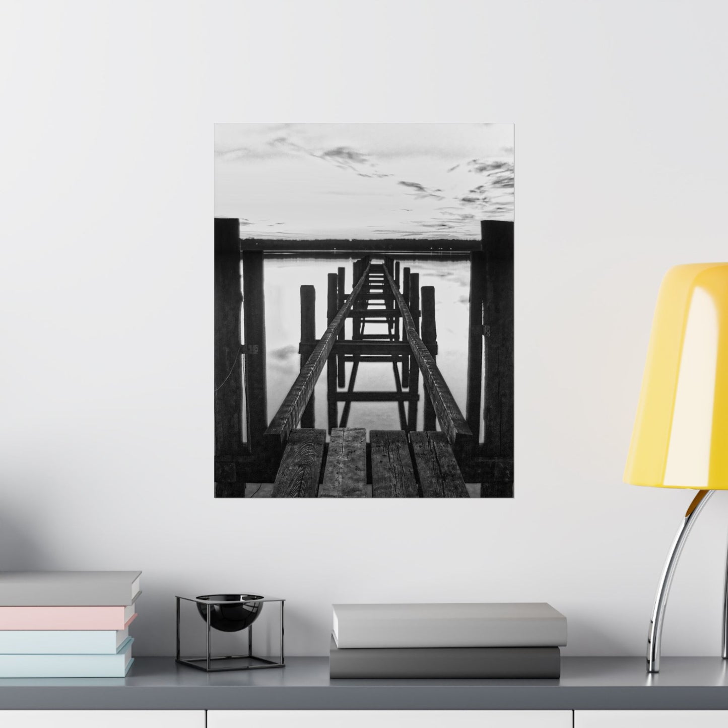 Framed black and white photograph of a dock on a wall above a shelf with books and a lamp.