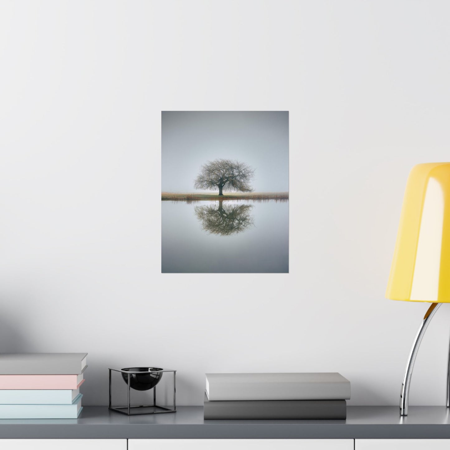 Framed photograph of a tree reflected in water on a wall above a desk with books and a lamp.