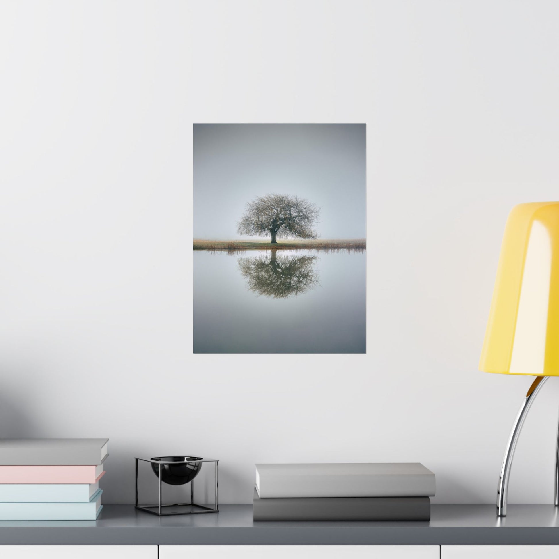 Framed photograph of a tree reflected in water on a wall above a desk with books and a lamp.