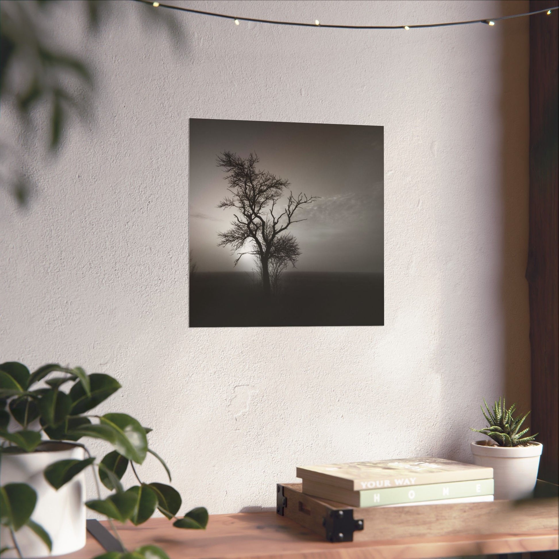 Art print of a lone tree on a white wall above a wooden desk with plants and books.