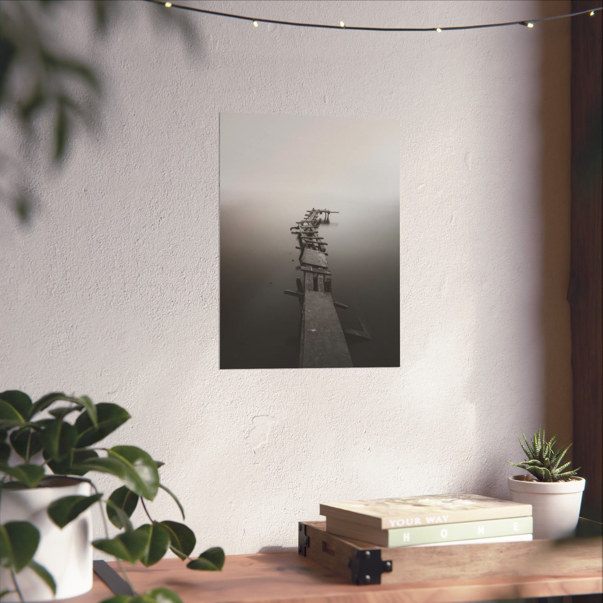 Framed black and white photograph of a dock on a wall above a wooden desk with plants and books.