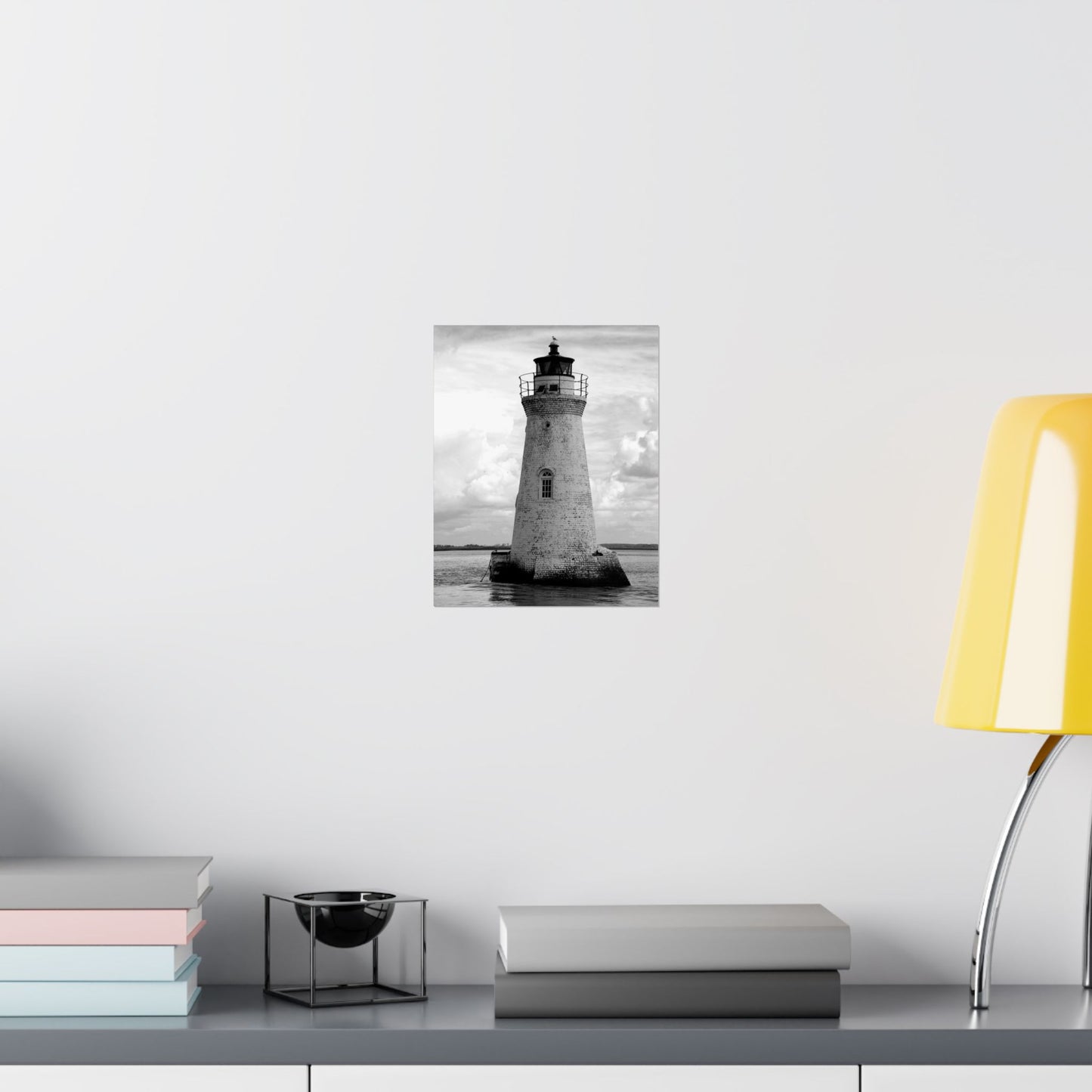 Framed black and white lighthouse print on a wall above a shelf with books and a lamp.