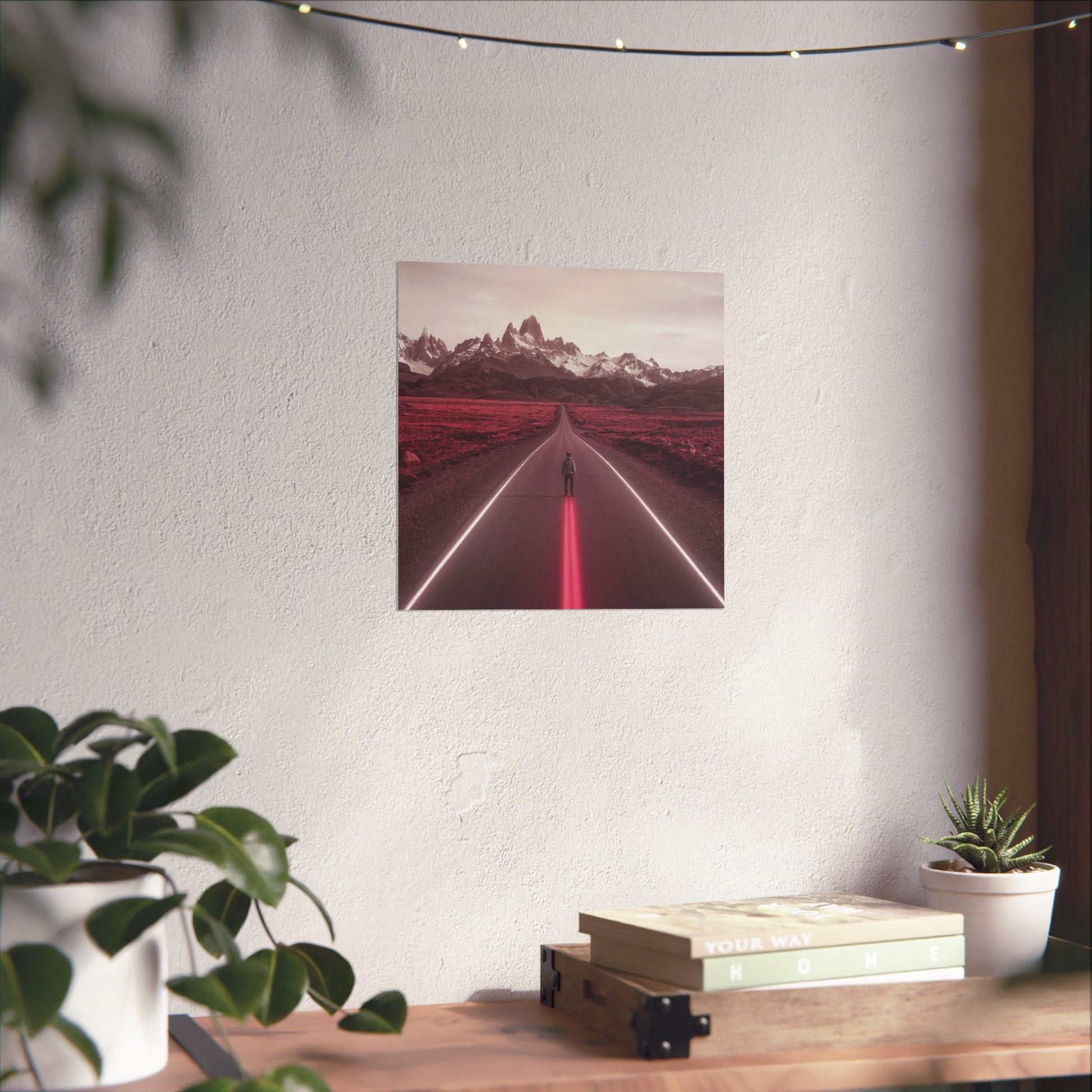 Framed artwork of a road with mountains on a white wall above a wooden desk with plants.