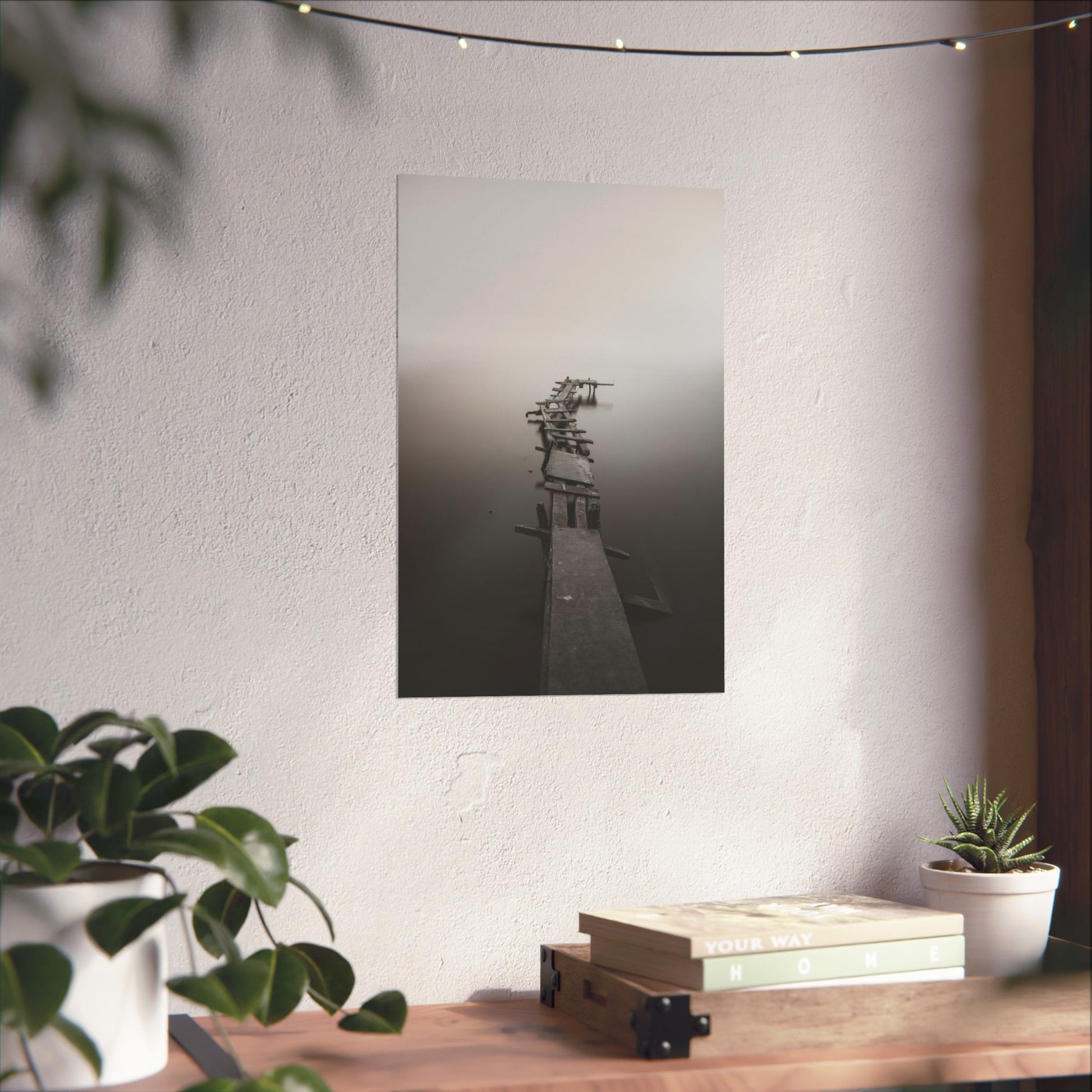 Framed black and white photograph of a dock on a wall above a wooden desk with plants and books.