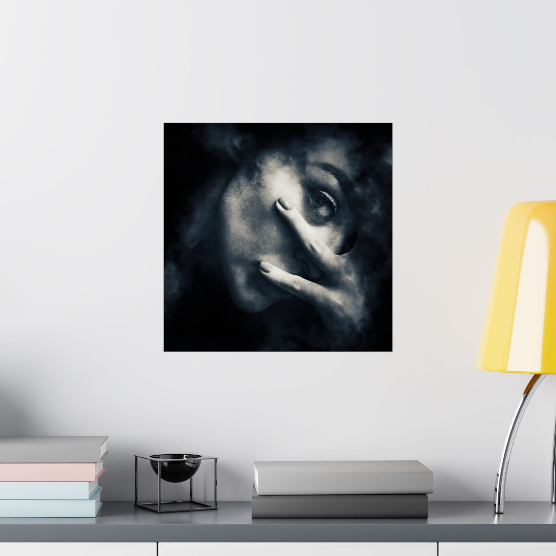 Framed artwork of a face with a hand over one eye on a wall above a desk with books and a lamp.