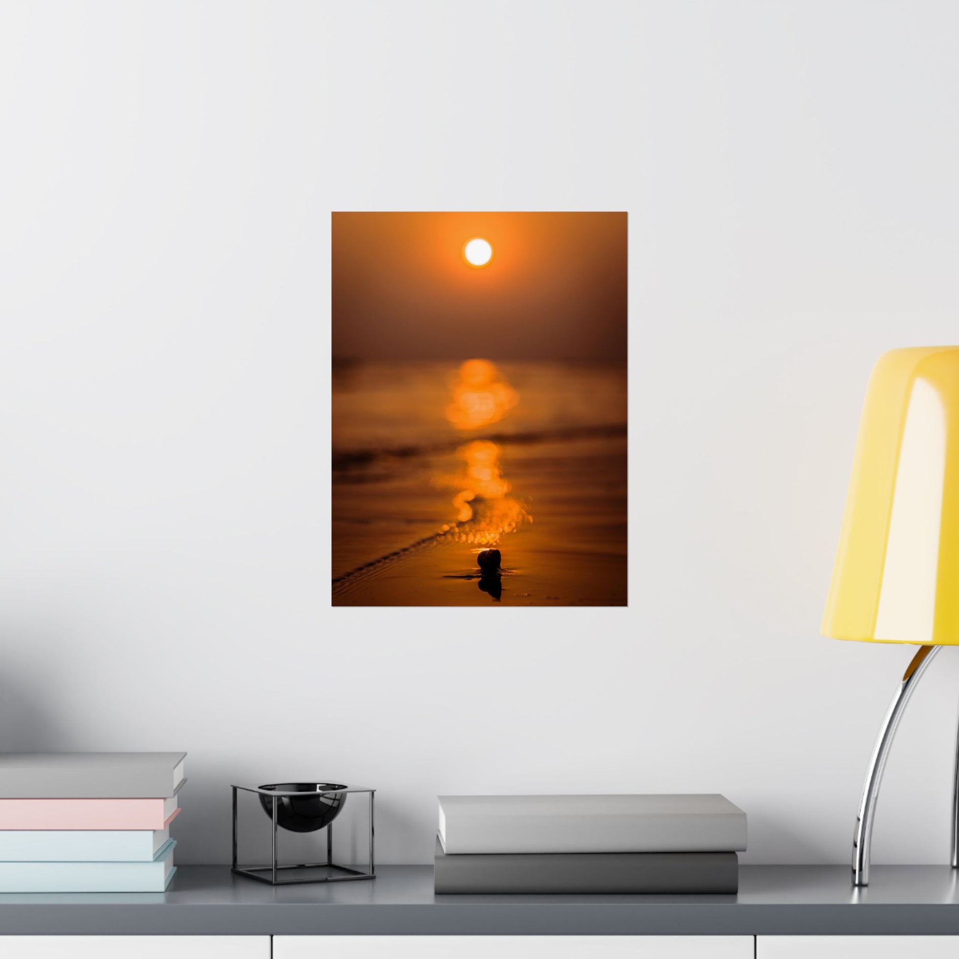 Sunset over water painting on a wall above a shelf with books and a lamp.