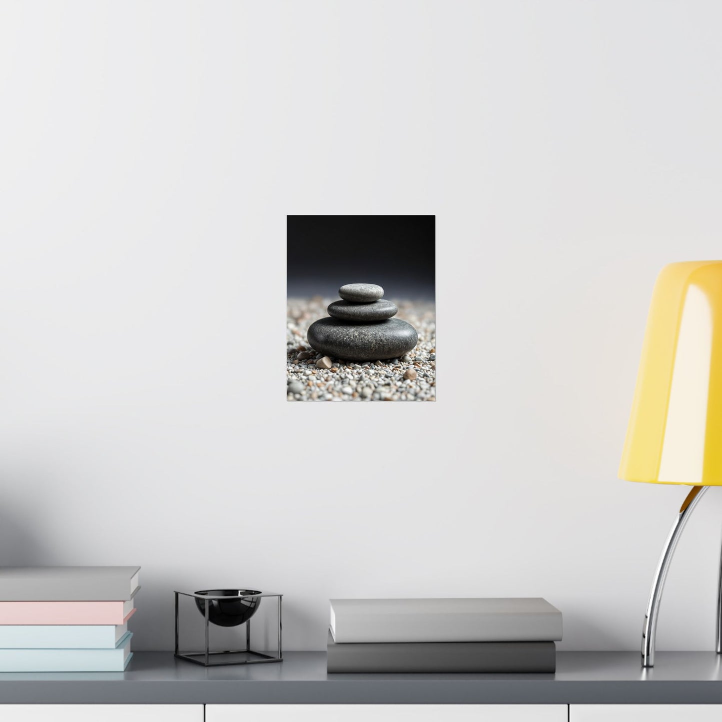 Wall art of stacked stones on pebbles with a yellow lamp and books on a shelf.