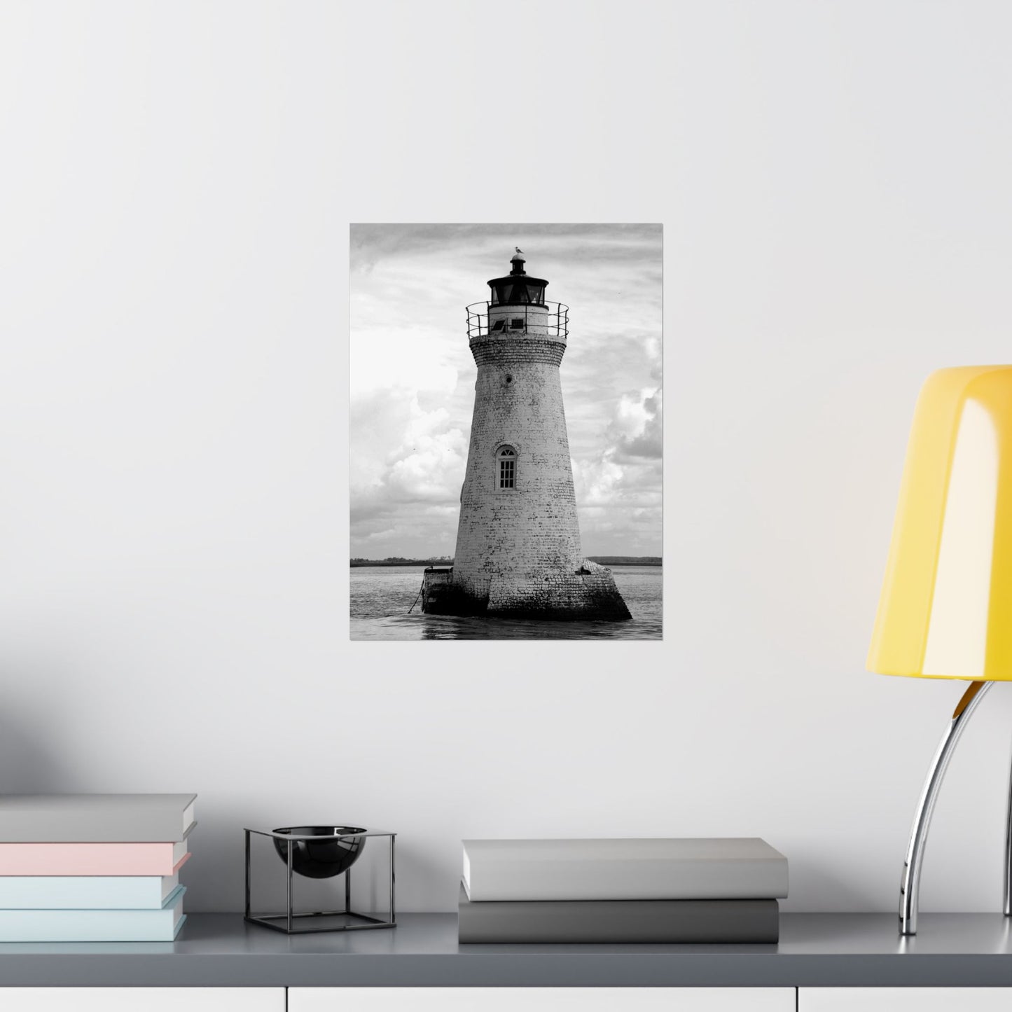Framed black and white lighthouse print on a wall above a desk with books and a lamp.