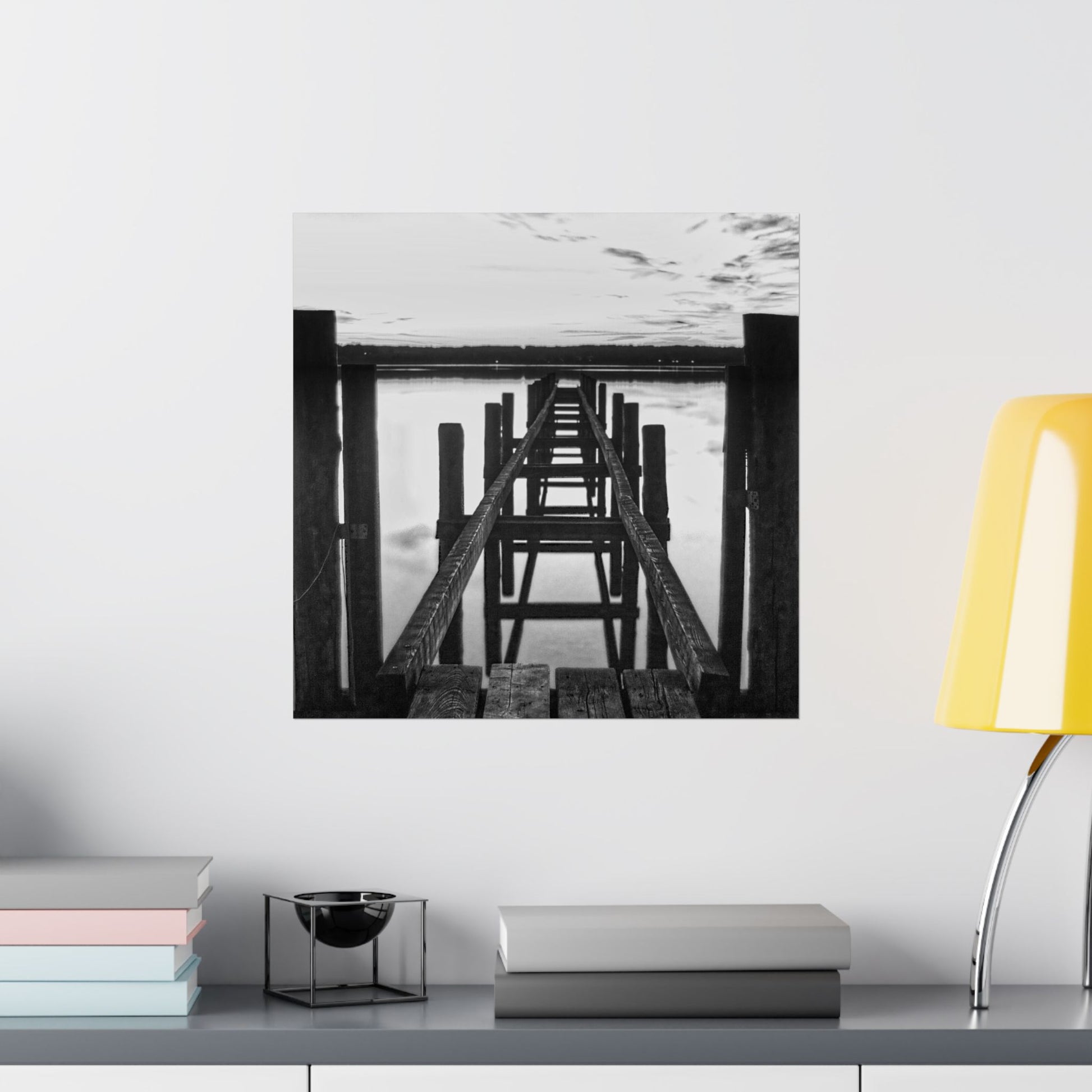 Framed black and white artwork of a dock on a wall above a shelf with books and decor.
