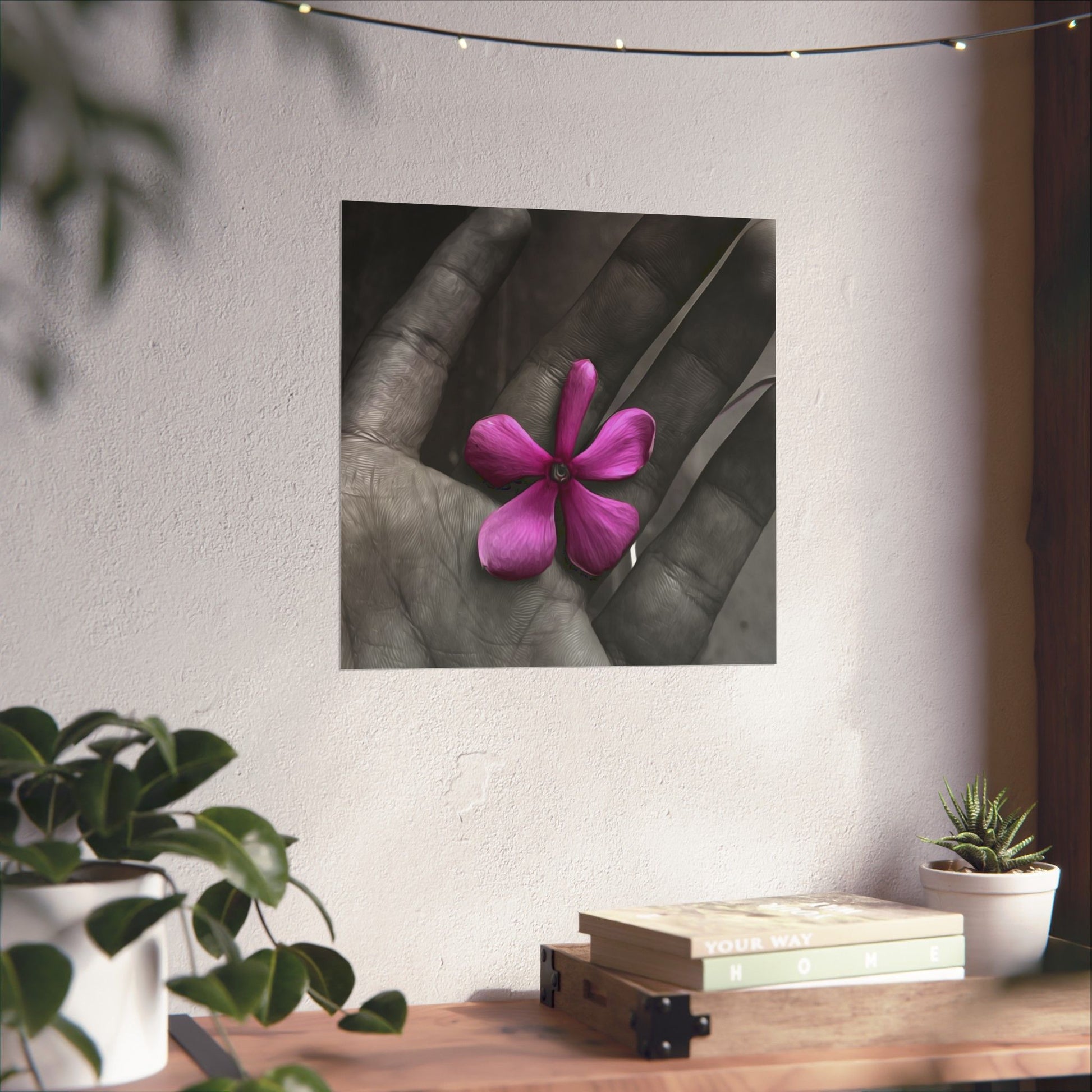 Framed artwork of a hand holding a pink flower on a wall above a wooden table with plants.