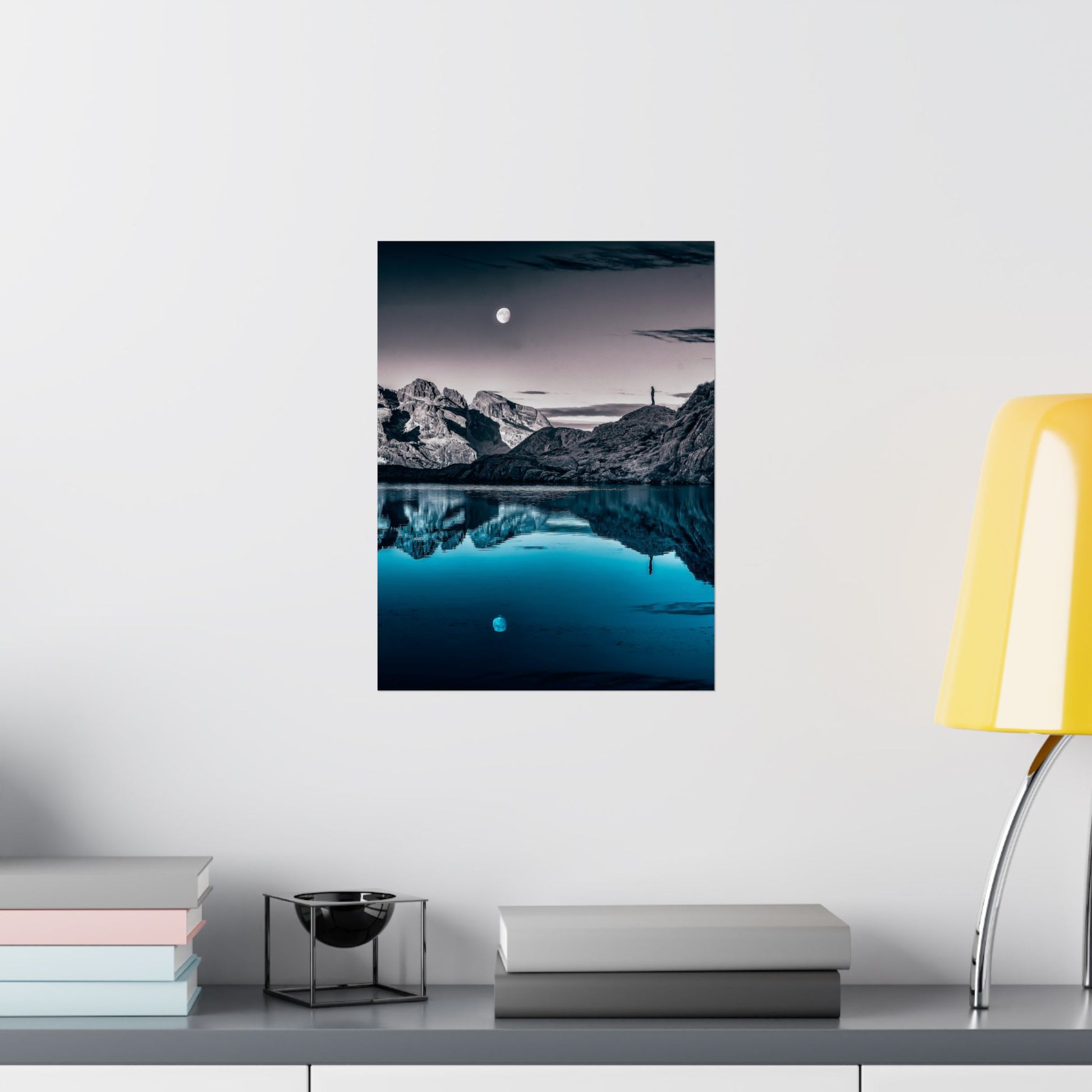 Framed mountain landscape print on a wall above a shelf with books and a lamp.