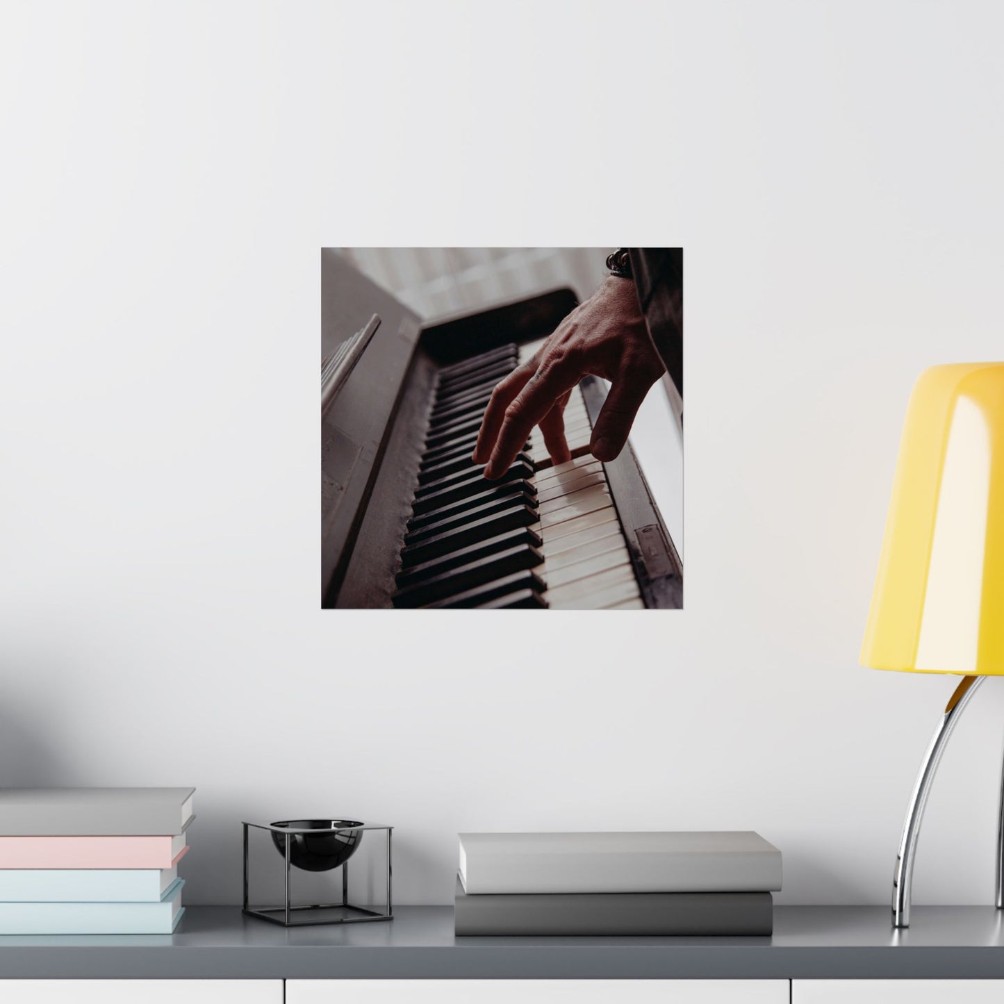 Framed picture of hands playing a piano on a wall above a desk with books and a lamp.