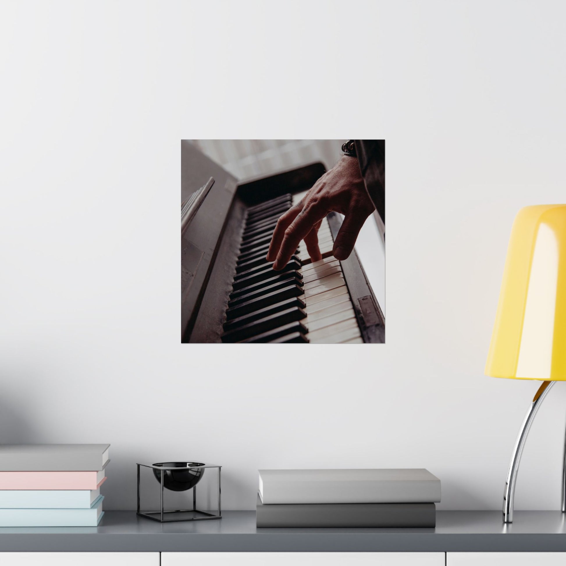 Framed picture of hands playing a piano on a wall above a desk with books and a lamp.