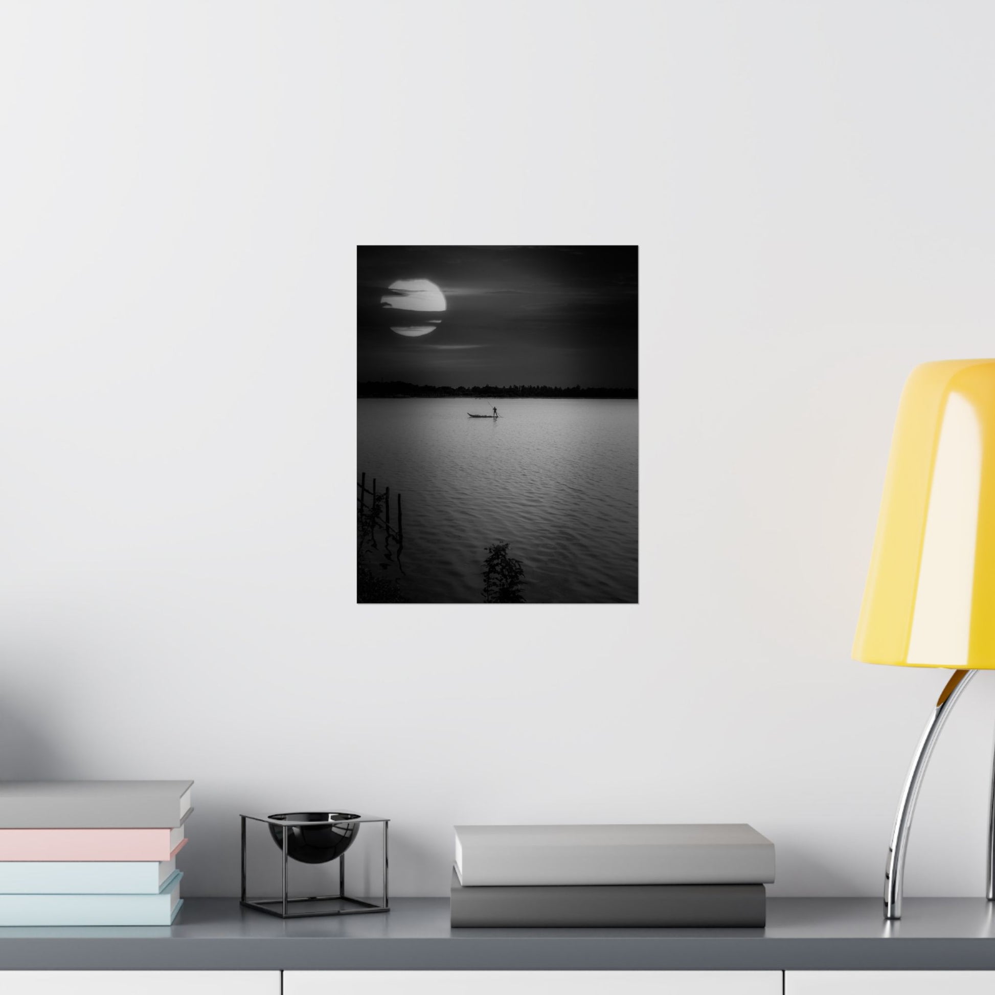 Framed black and white artwork of a moonlit lake on a wall above a desk with books and a lamp.