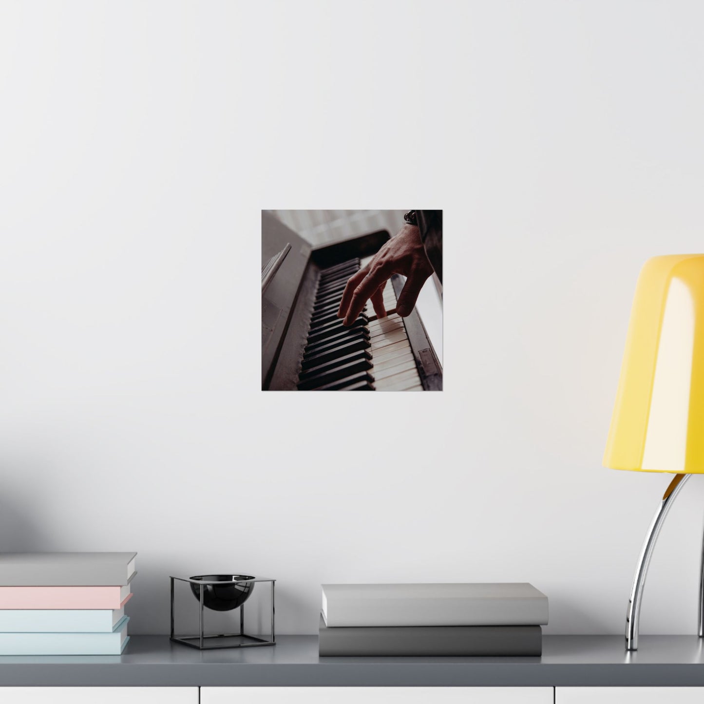 Framed picture of hands playing piano on a wall above a desk with books and a lamp.